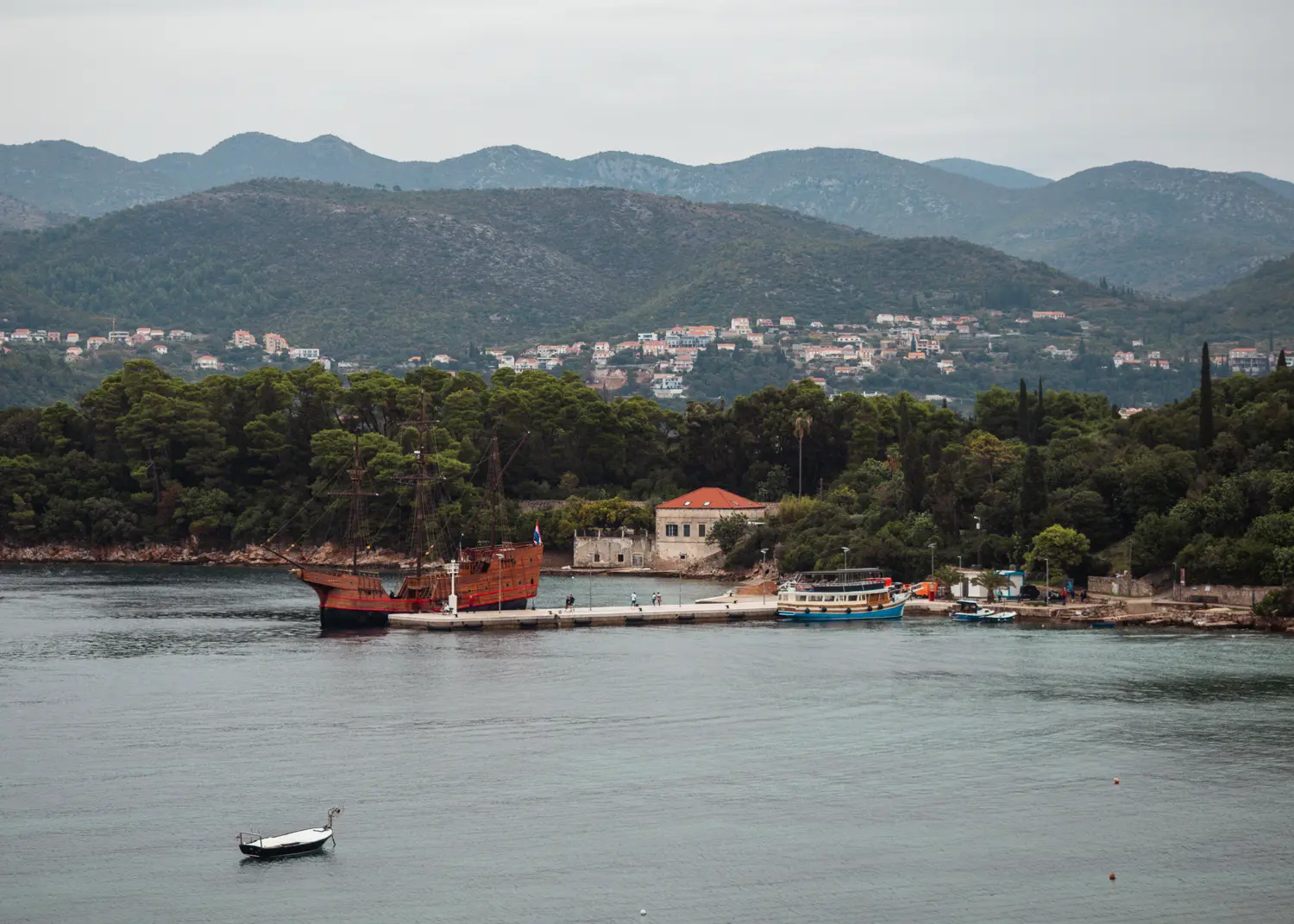 Brown wooden ship docked at Kolocep Harbor with forest and mountains in the background seen from the balcony of my room at TUI BLUE Kalamota Island Resort.
