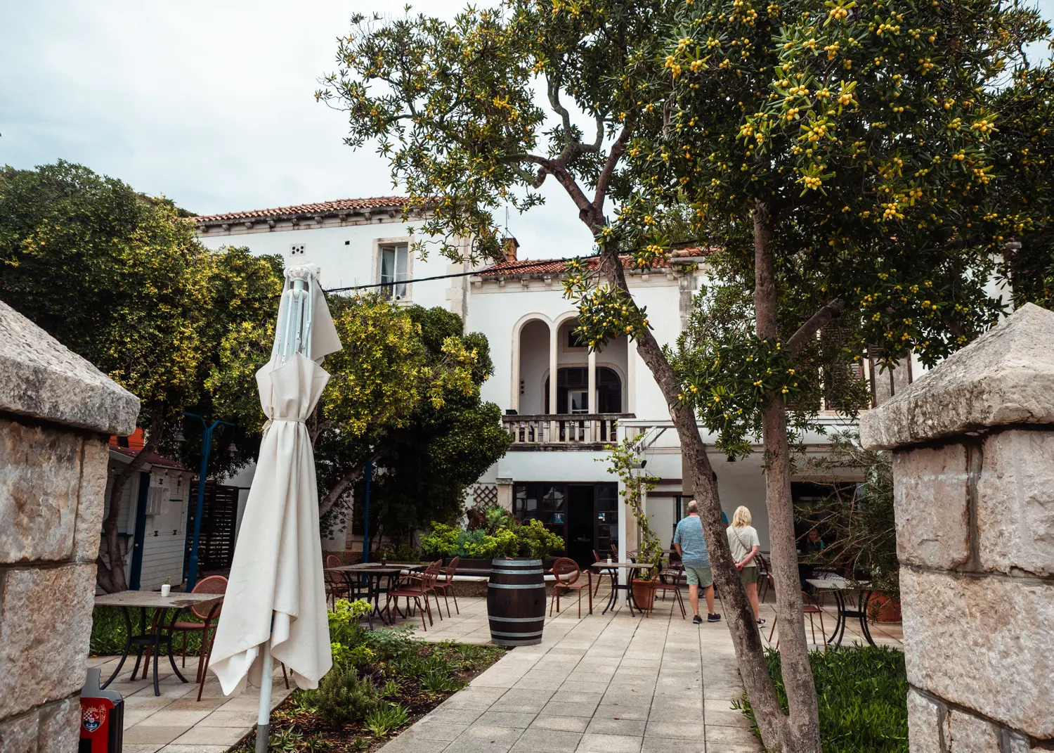 Looking though a stone gate on Culinarium Restaurant set in a white Mediterranean style house with green trees in front, close to TUI BLUE Kalamota Island Resort.