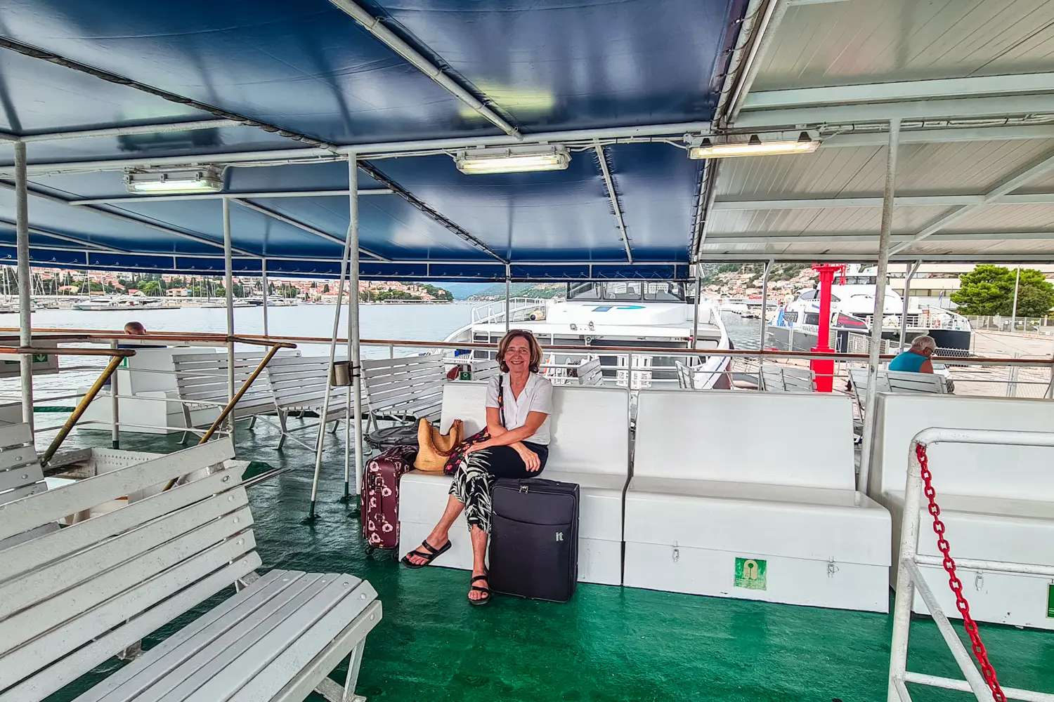 Woman wearing a white shirt and black pants, sitting next to one black and one burgundy suitcase in the middle of a ferry with green floor and white seating in the way to TUI BLUE Kalamota Island Resort on Kolocep, Dubrovnik, Croatia