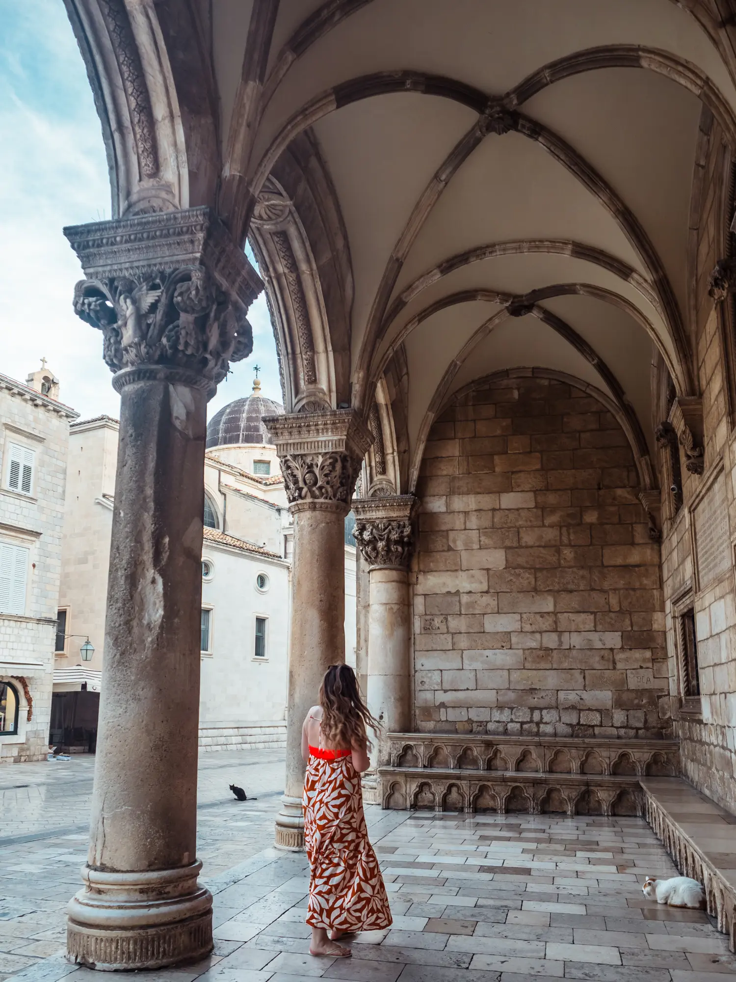 Woman with long dark blonde hair wearing a long orange and white dress under the large pillars and vaulted ceiling of Rector's Palace during a walking tour in Old Town Dubrovnik.