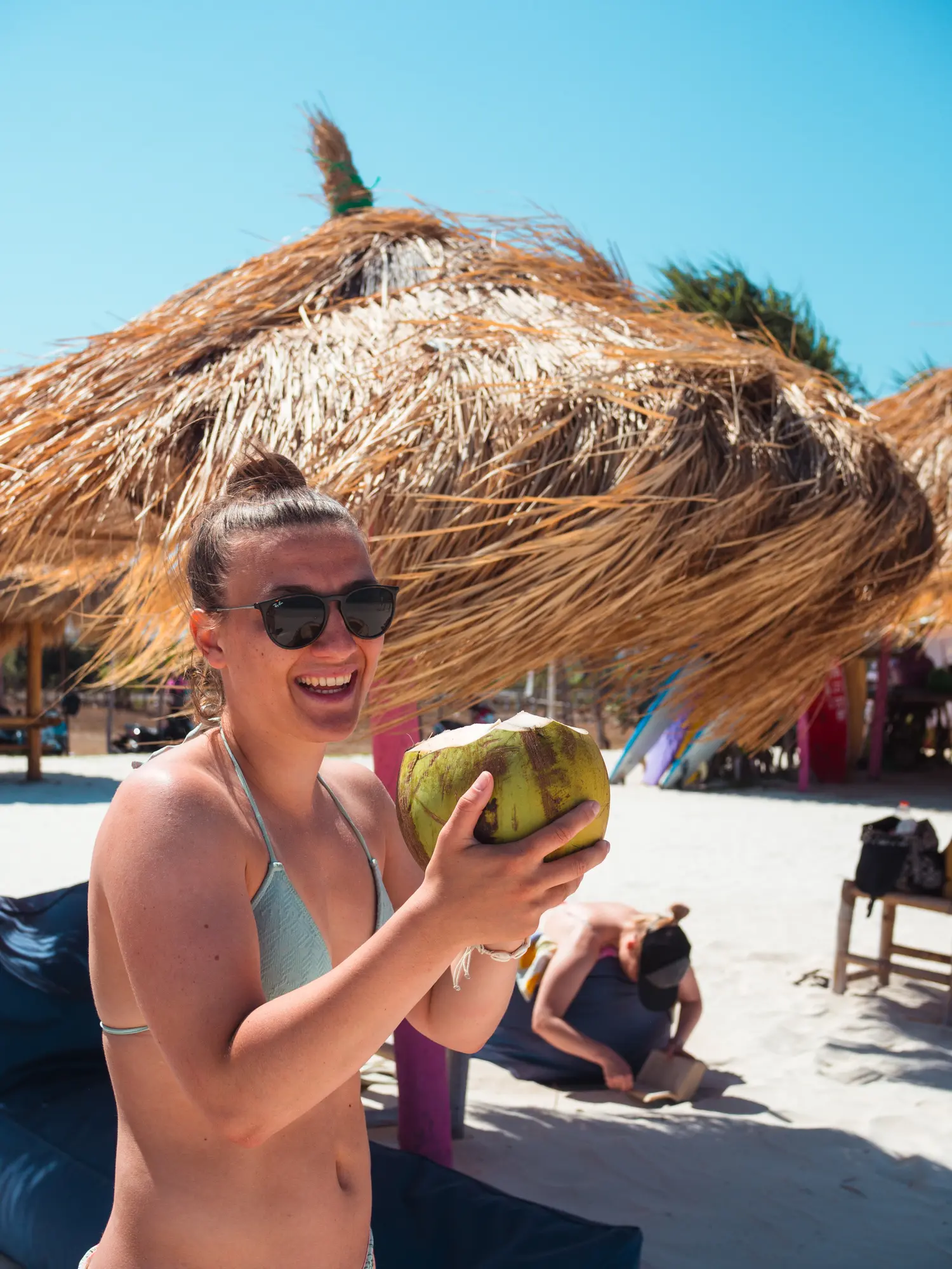 woman wearing a blue bikini and sunglasses smiling to the camera while holding a green coconut with both hands between surf sessions during a Lombok surf camp.