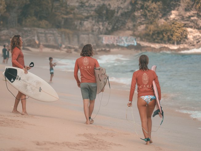 3 surfers wearing red rash guards, holding surfboards, walking along the beach during a surf and yoga retreat in Bali.