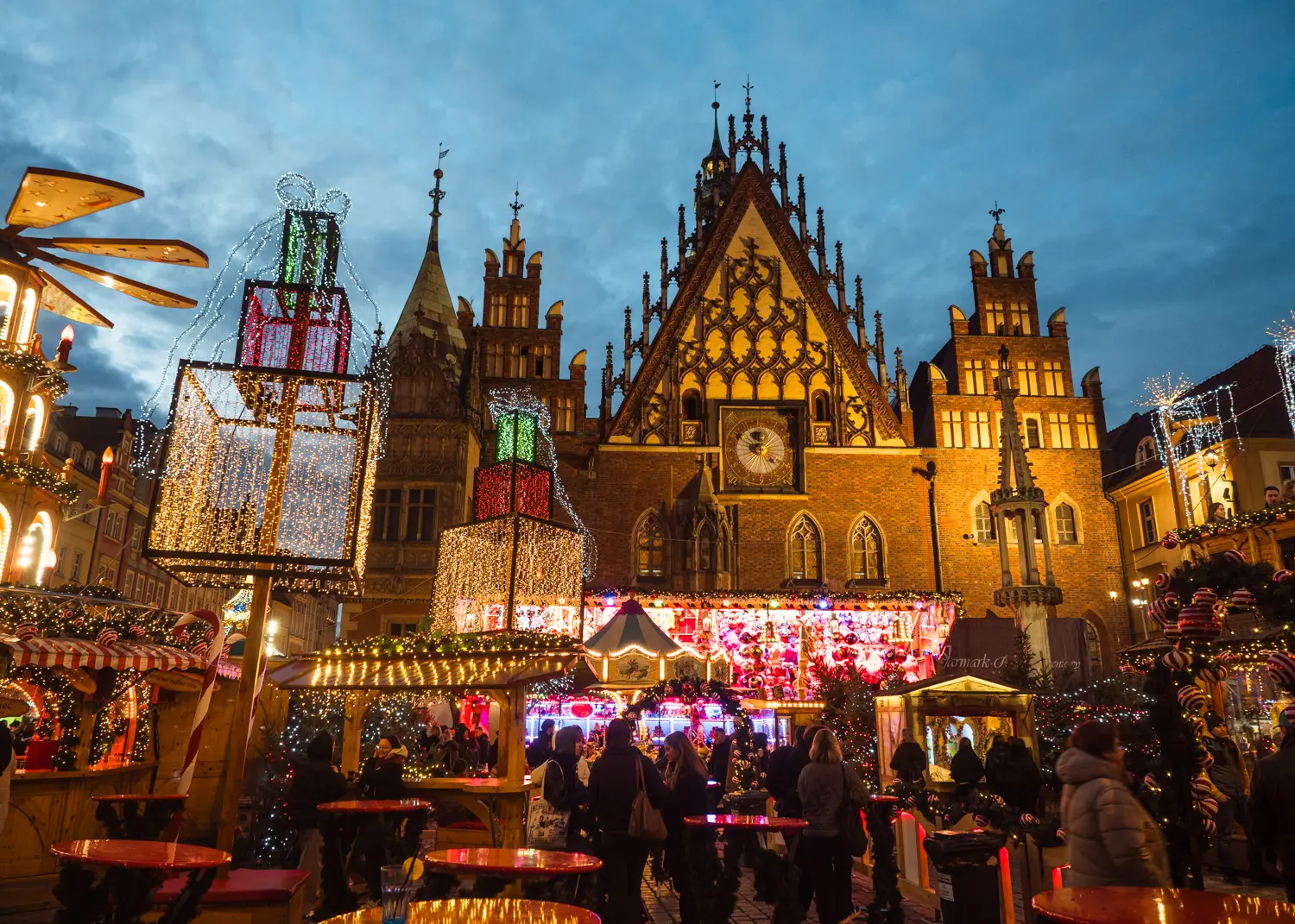 People standing around red tables surrounded by light and decorations in from of the large Gothic brick Town Hall at Wroclaw Christmas Market.