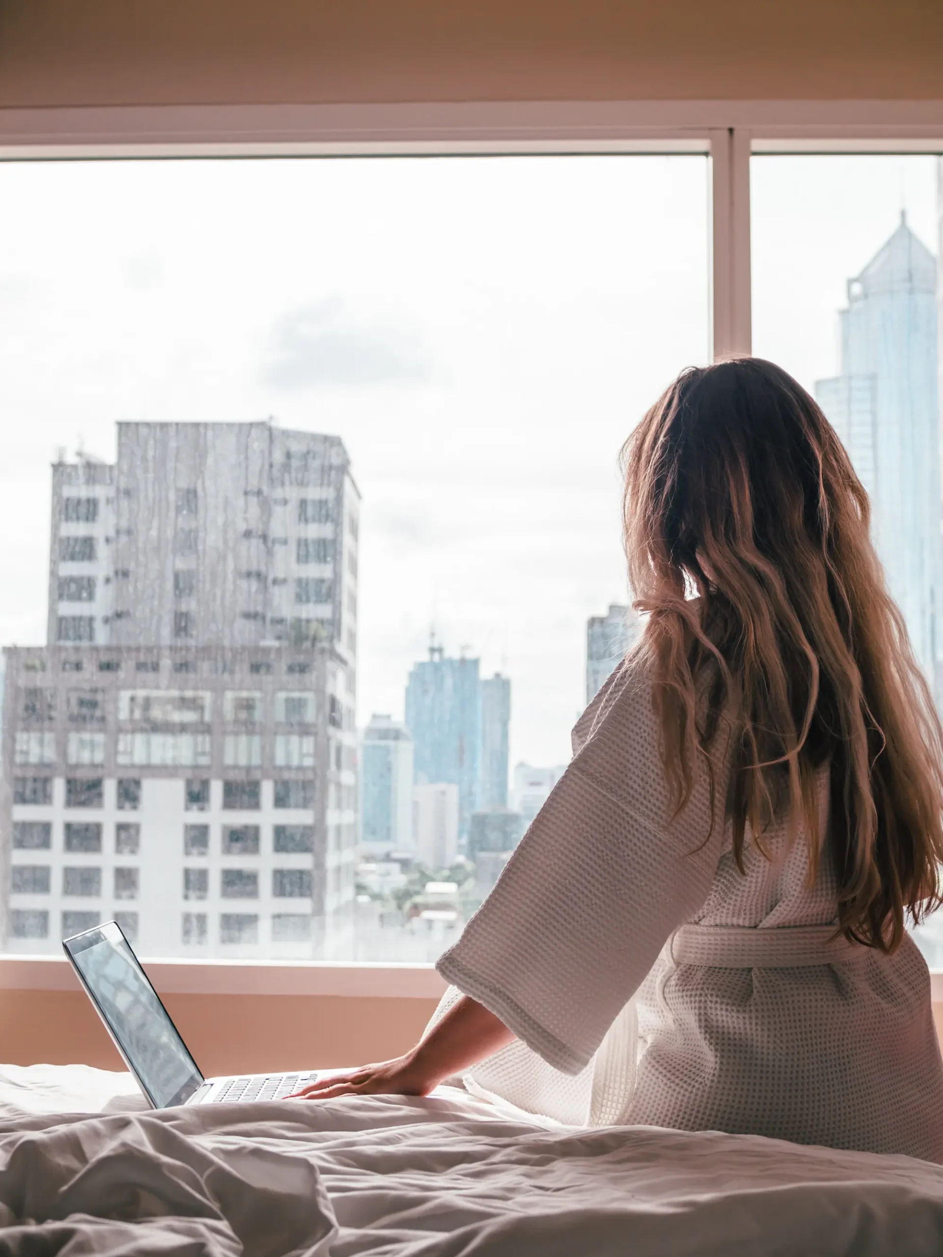 Woman with long dark blonde hair, wearing a white robe, sitting on a bed with a laptop looking out a large window on skyscrapers in a hotel while working as a content creator for free accommodation while traveling. 