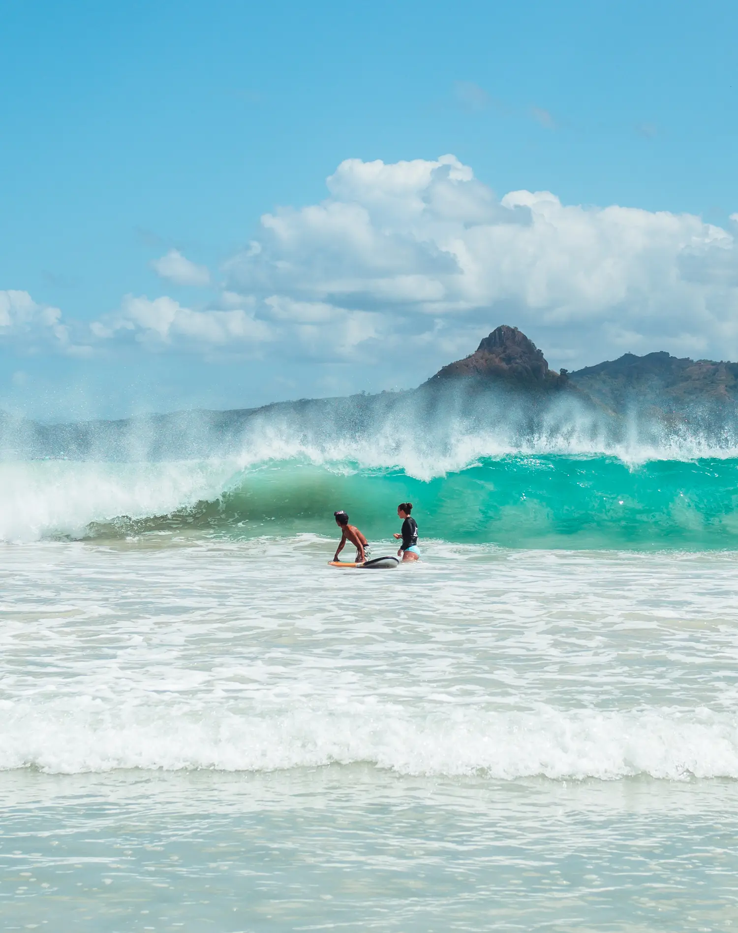 Beginner surfer and instructor standing in the turquoise ocean with a big wave breaking behind them, during a surf camp in Lombok.