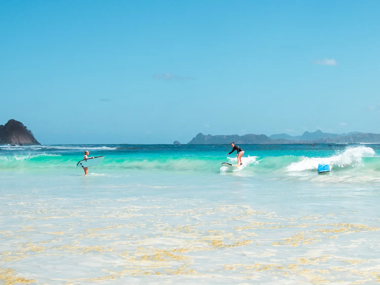 Beginner surfer standing up on a small wave with another surfer looking in the turquoise shallow ocean of Selong Belanak Beach, as part of one of the best surf camps in Lombok.