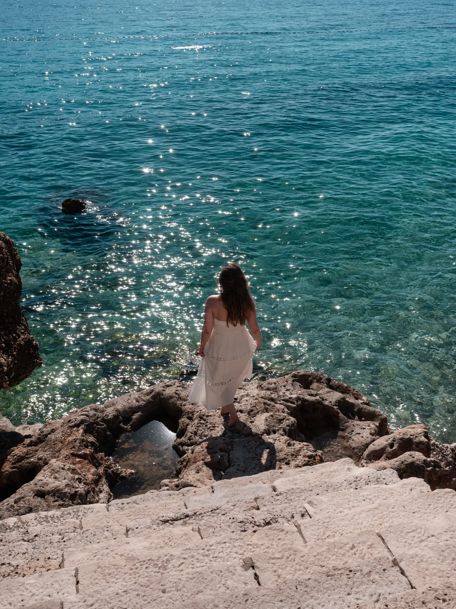 Woman with brown hair, wearing a white dress, walking down a stone staircase towards somw rocks by the glistening ocean at Banje Beach in the Ploce neighborhood, where to stay in Dubrovnik.