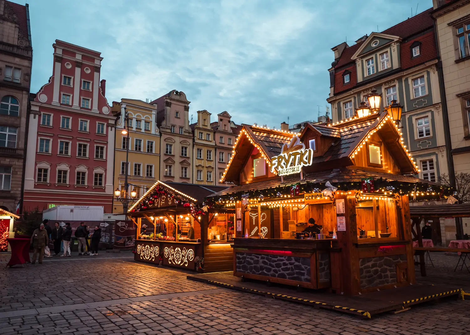 Two small wooden houses/stalls decorated with lights in the middle of the cobbled square surrounded by tall colorful town houses at Wroclaw Christmas Market.