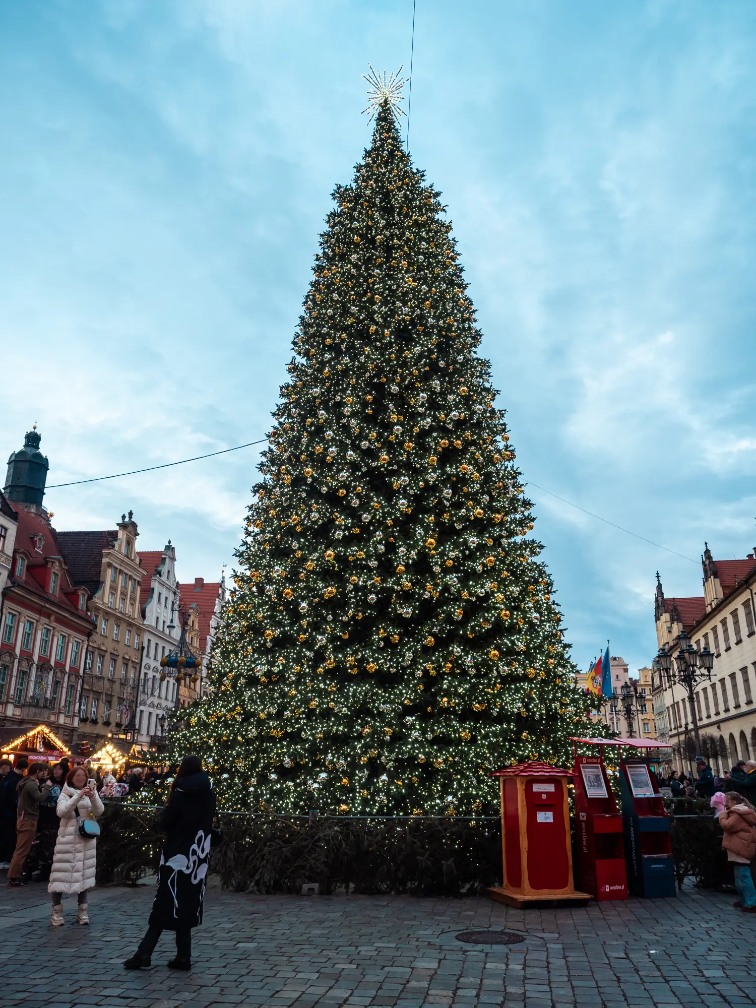 Massive Christmas tree evenly decorated lights and baubles with a red mailbox and people taking photos in front at Wroclaw Christmas Market.