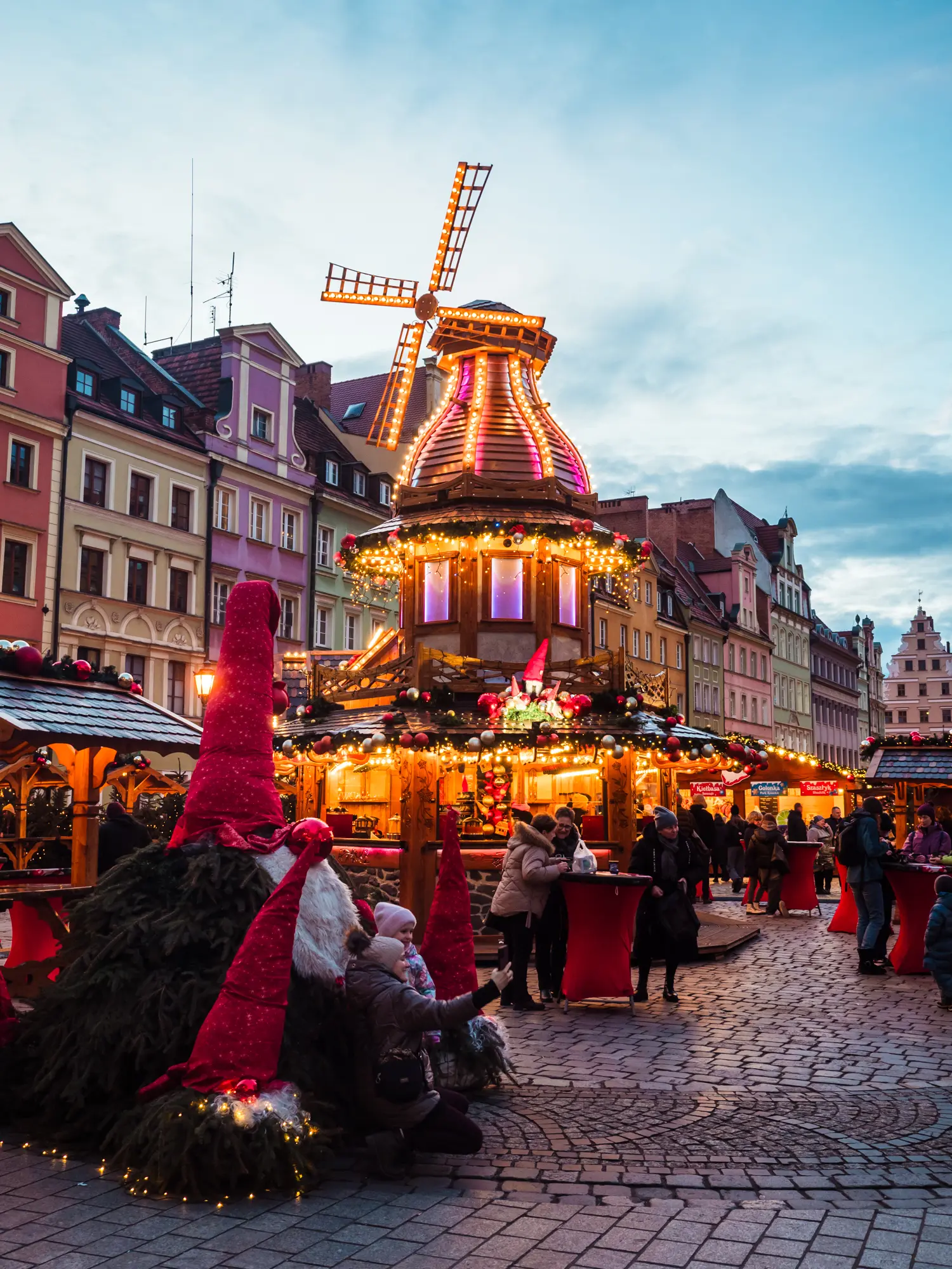 View of a wooden windmill will lights in many colors, Santas made from pine trees in the foreground and old town houses in the background and Wroclaw Christmas Market.