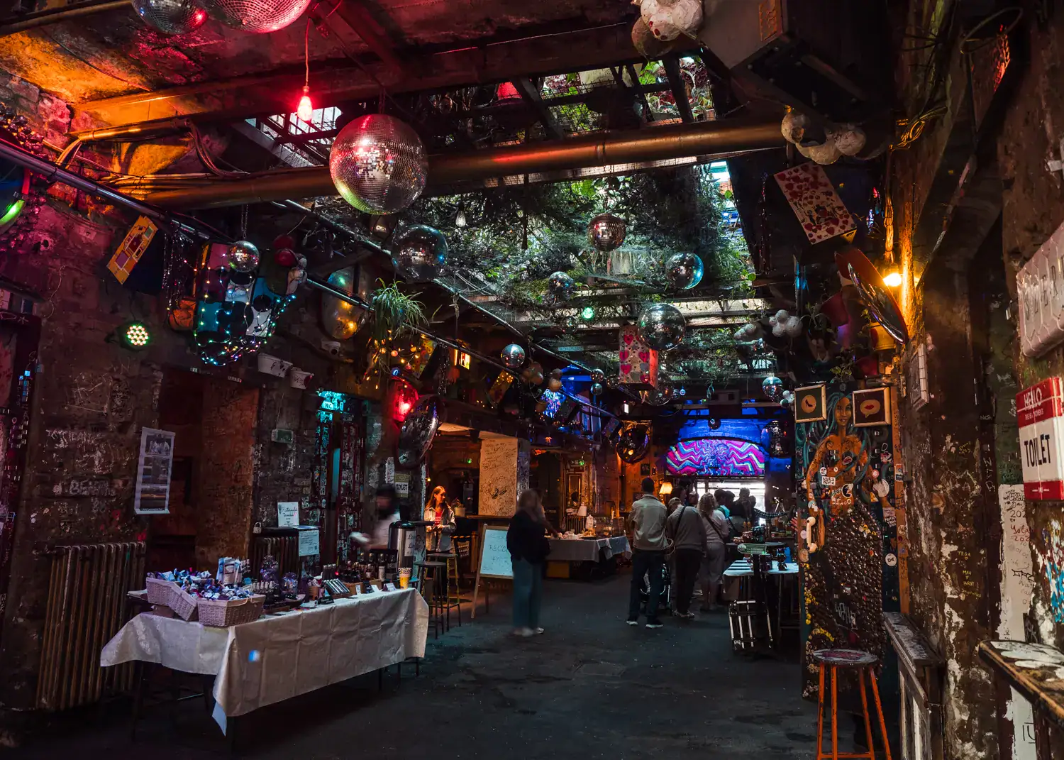 View inside the quirky and dark Szimpla Kert ruin bar in Budapest during their popular Sunday Farmers' Market. Tables on each side, a few people walking around, disco balls and greenery in the ceiling, fairy lights.