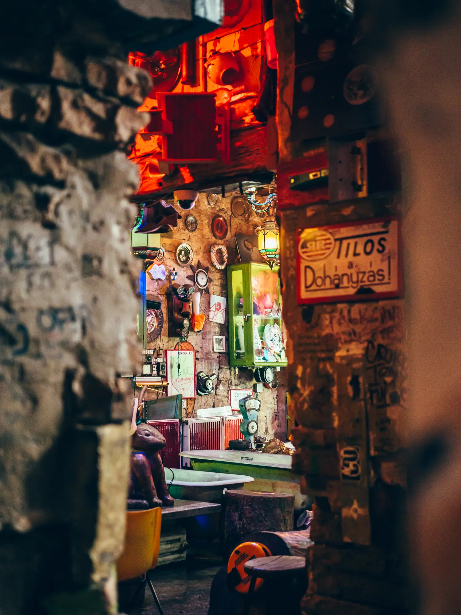 Looking through the stone entrance to one of the bars inside Szimpla Kert in Budapest. Bathtubs, green glass display cases on the stone walls, a metal hare, lots of little pictures and trinkets.
