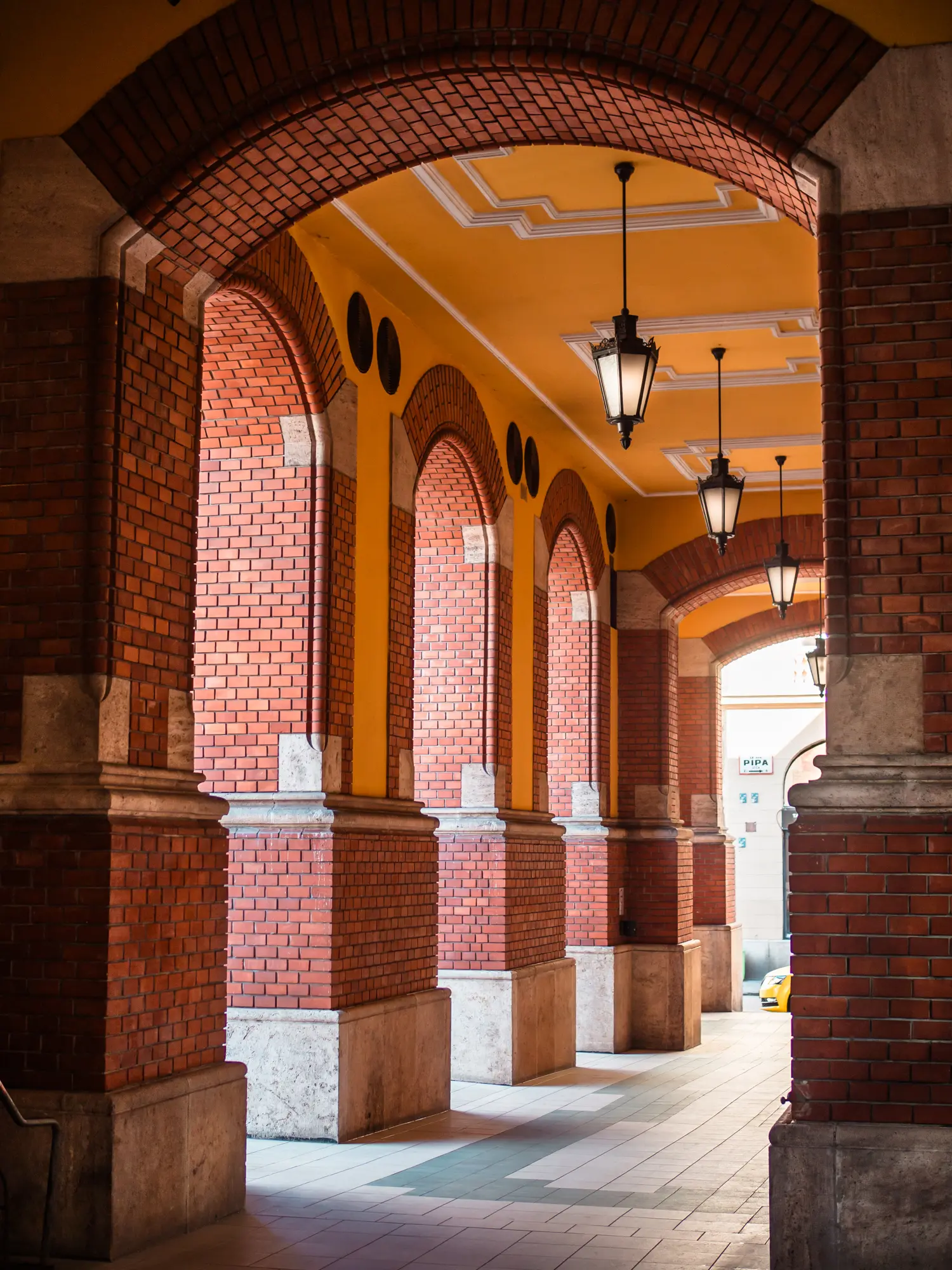 Covered outside walkway with red brick arches and yellow decorative ceiling with four black metal lights at the Central Market Hall in Budapest.