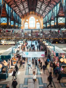 Woman in a white coat standing in the middle of Budapest Central Market Hall with lots of people walking around her, seen from the upper level of the metal building with vaulted ceilings.