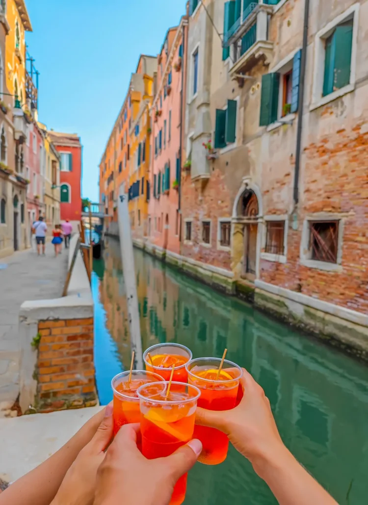 Four hands holding red drinks in front of a green canal in Venice with brick buildings on each side while hosting a group trips to boost my content creator income.