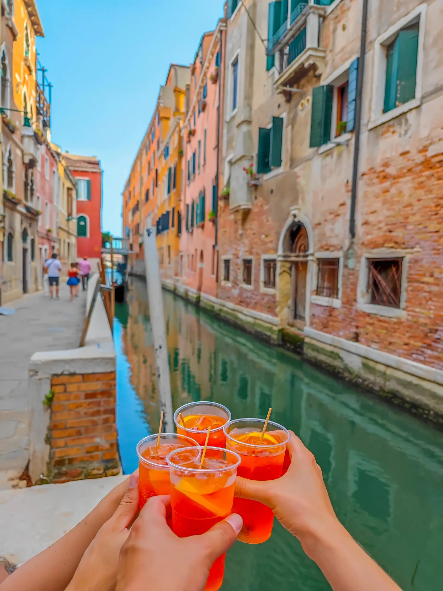 Four hands holding red drinks in front of a green canal in Venice with brick buildings on each side while hosting a group trips to boost my content creator income.