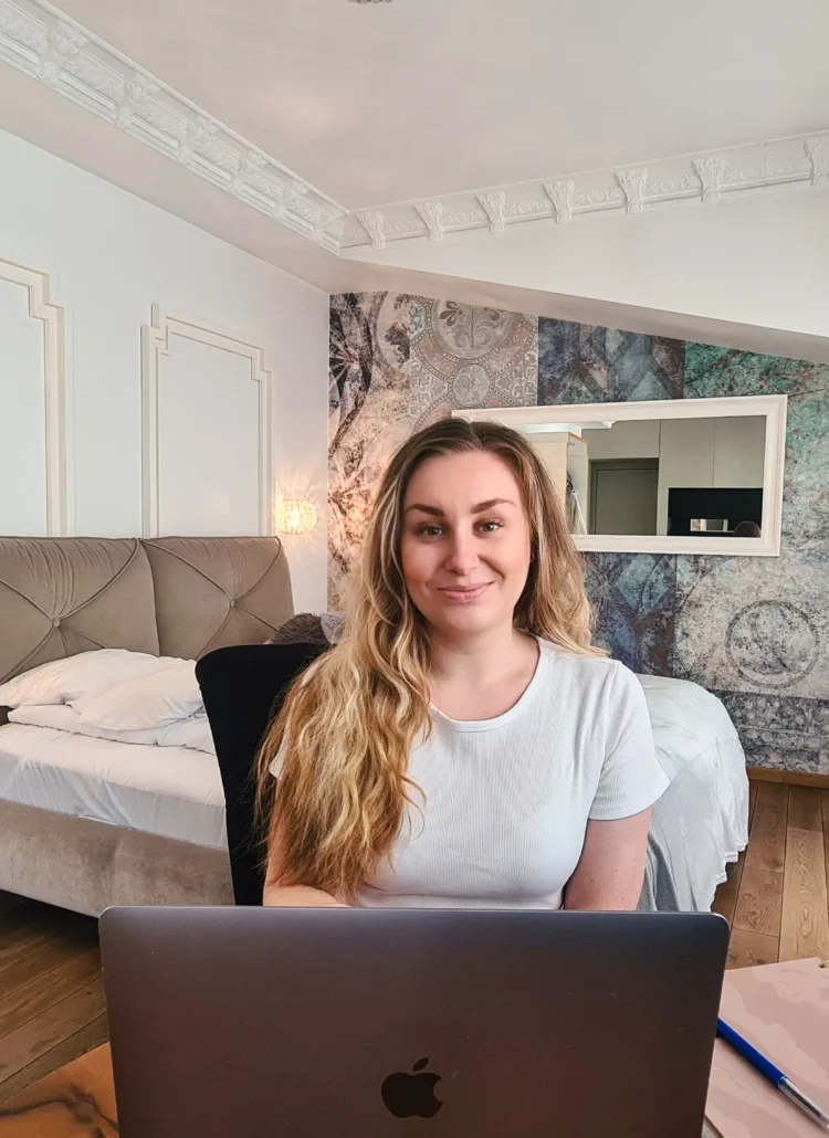 Woman with long dark blonde hair, wearing a white t-shirt, sitting behind a silver laptop in a grand apartment working on growing travel blog traffic to get accepted by Mediavine ads.