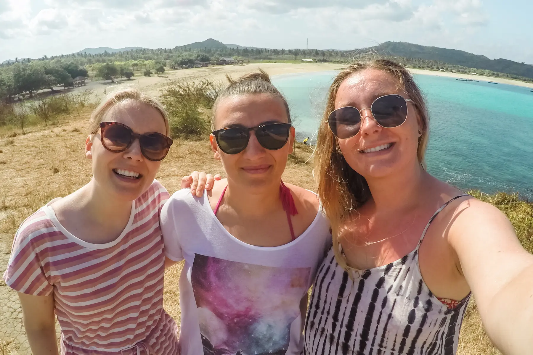 Three girls, wearing sunglasses, smiling to the camera while taking a selfie with a beach on the background during a group trip hosted by a creator.