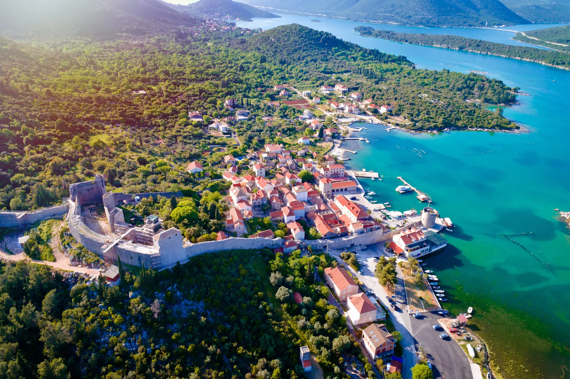 Aerial view of Mali Ston Old Town with a long stone wall and houses with orange roofs by the turquoise ocean, the perfect stop on our way from Split to Dubrovnik.