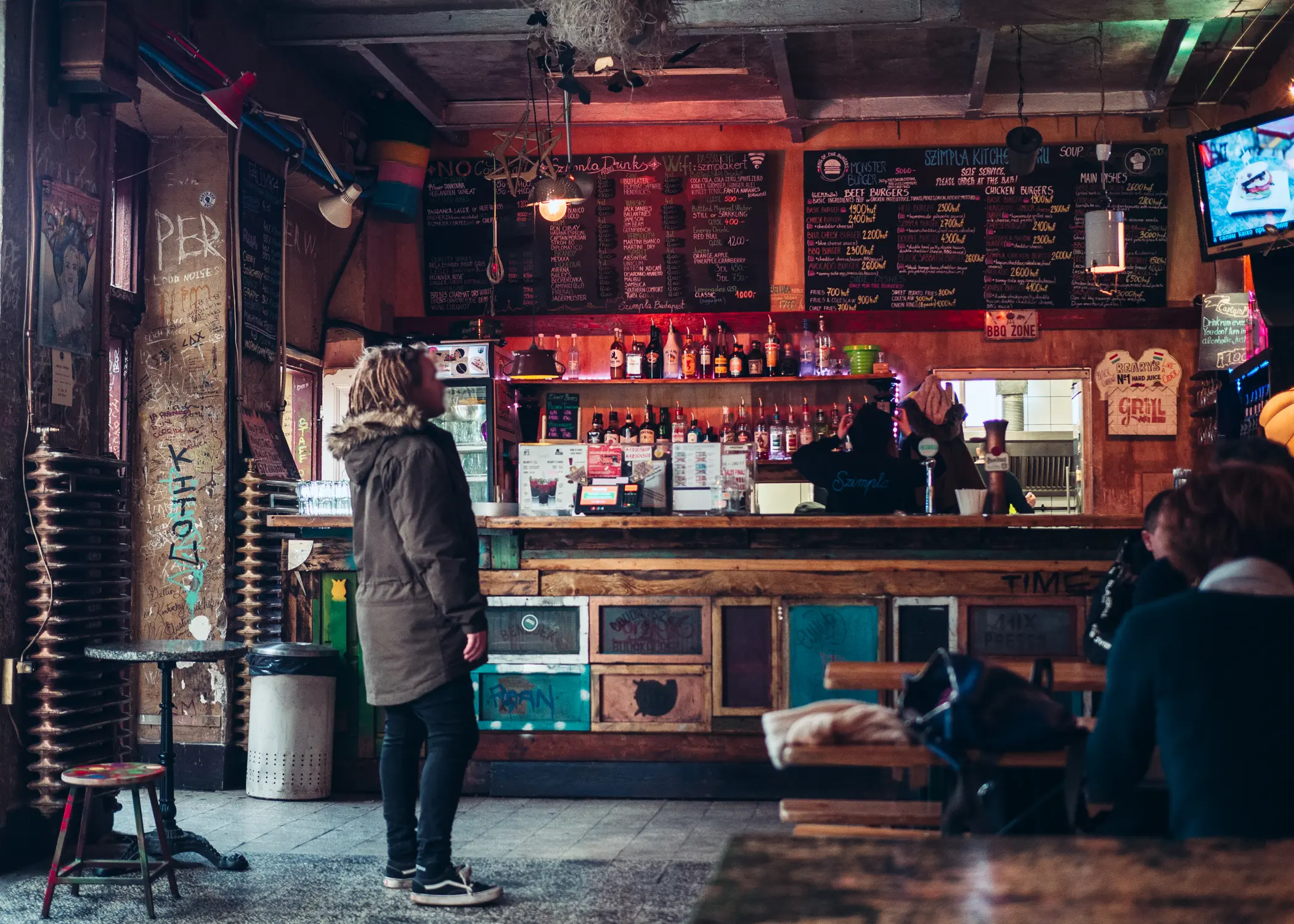 Man wearing black pants and a grey winter coat, standing in the middle of a bar in Szimpla Kert, looking at a tv on the wall.