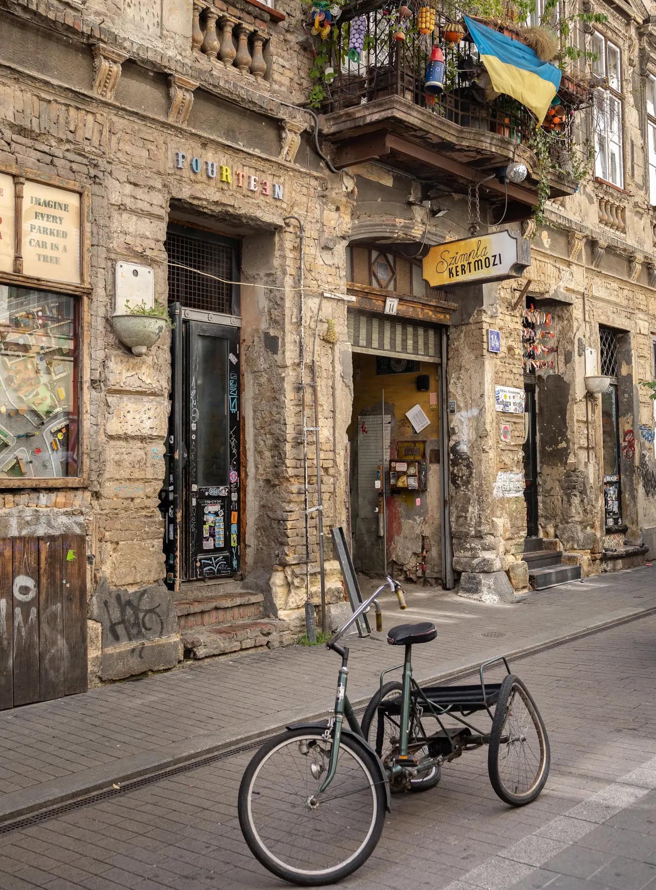 Old black tricycle in the street outside the entrance to Szimpla Kert ruin bar in a tired beige stone building with a yellow sign.