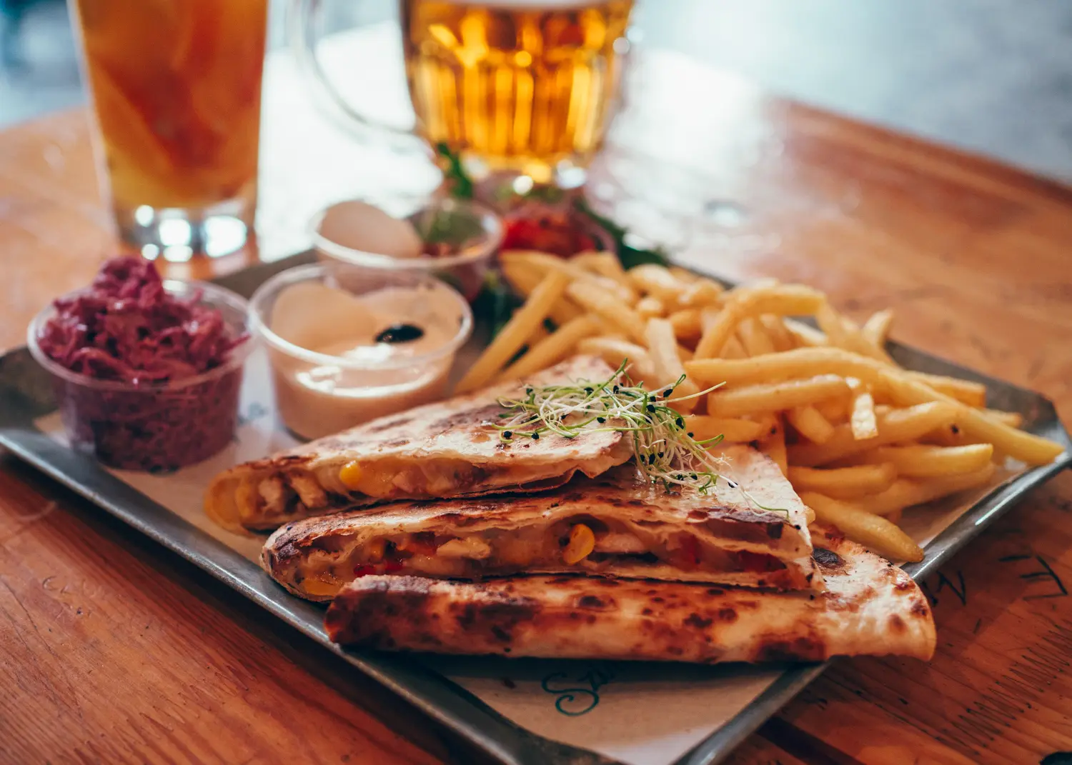 Metal tray with three pieces of quesadilla, French fries, coleslaw and a white dip, with a beer and iced tea in the background, in Szimpla Kart, Budapest.