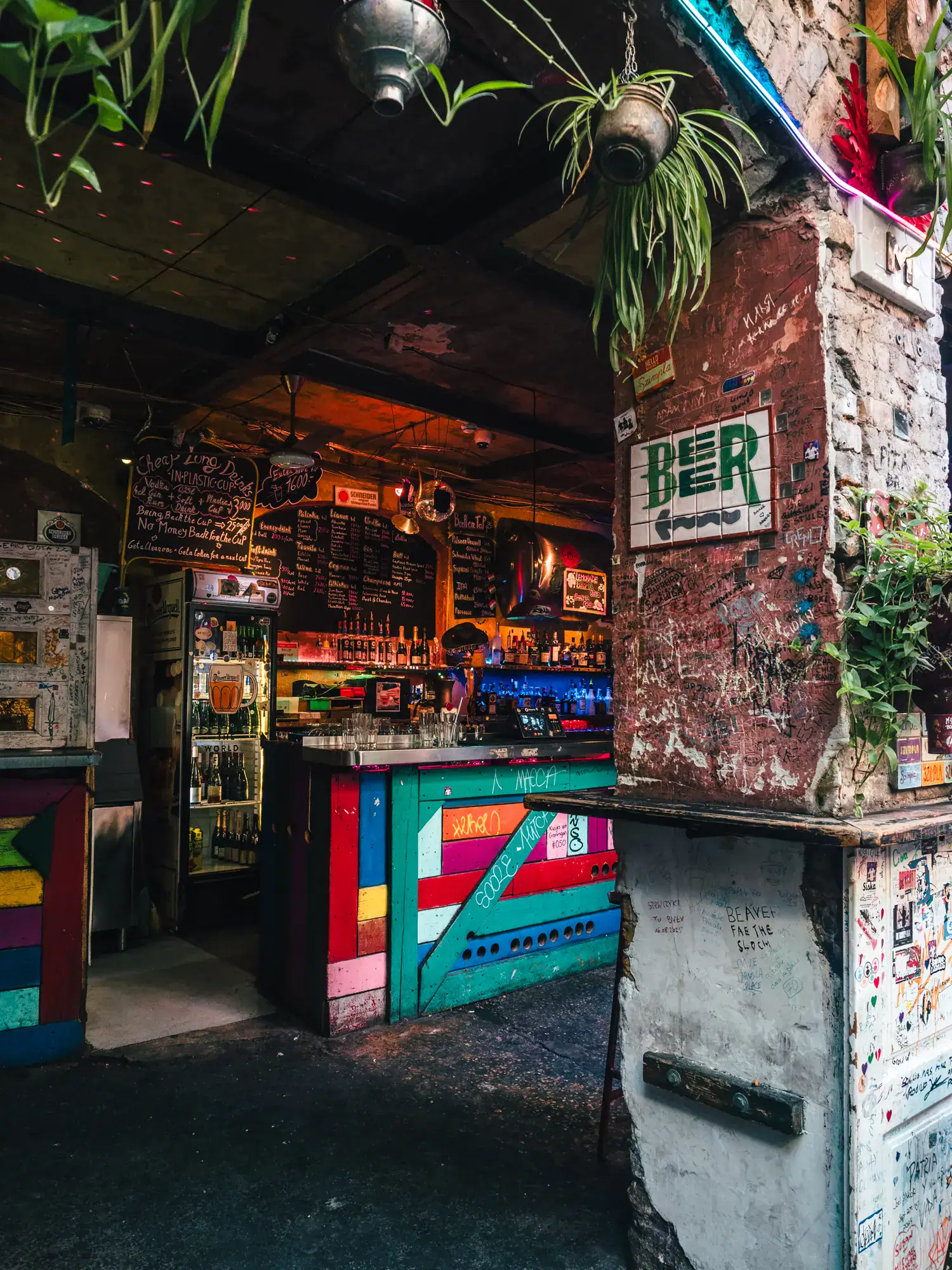Looking past a brick pillar into a colorful bar, beer sign on the pillar, lots of bottles on the wall behind, plants hanging above, at Szimpla Kert in Budapest.