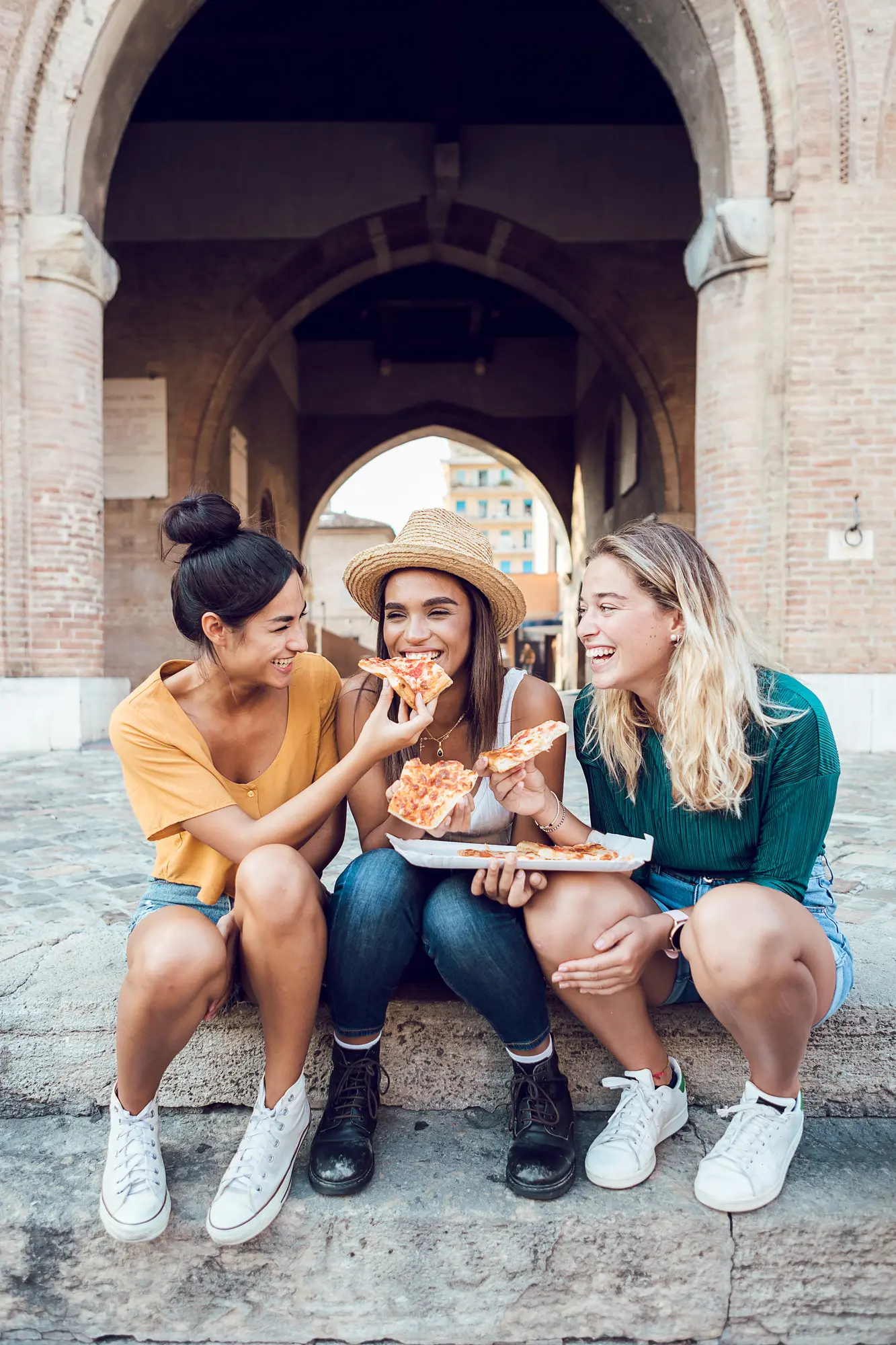 Three girls sitting on stone steps in front of an arch in Italy eating pizza during a group trip, a great way for creators to boost their income.