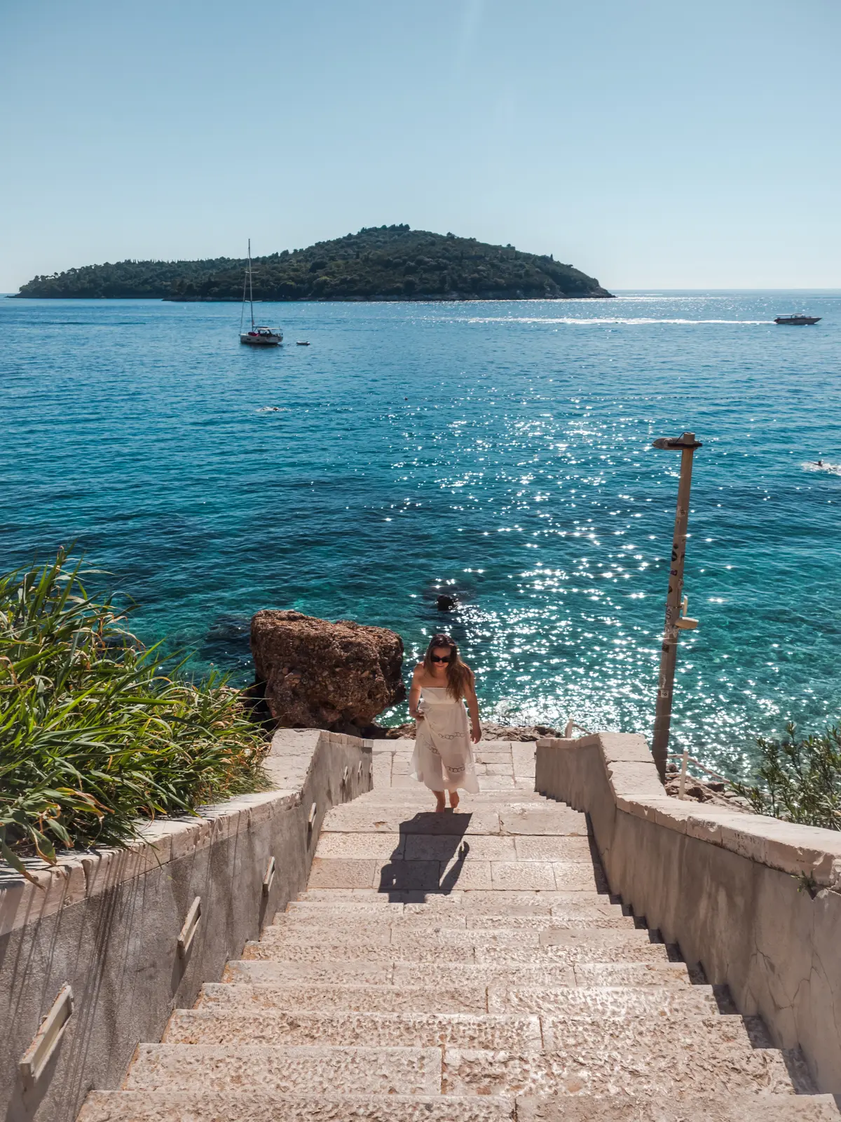 Woman with brown hair, wearing a white dress, walking up a stone staircase by the glistening ocean at Banje Beach, with a small island in the background, during an all-inclusive holiday in Croatia.