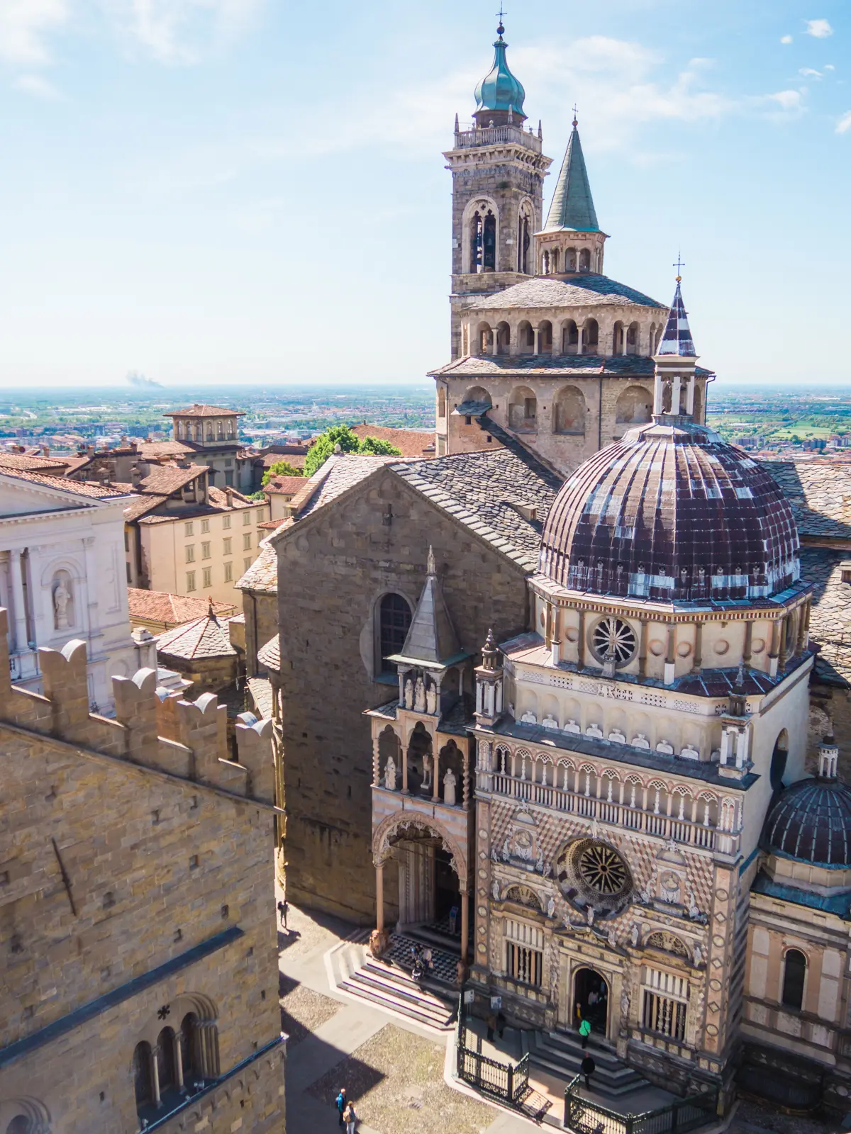 View from above of Basilica di Santa Maria Maggiore, with an ornate facade and dome with a tall spire in the background on a sunny day in Bergamo Citta Alta, Italy.