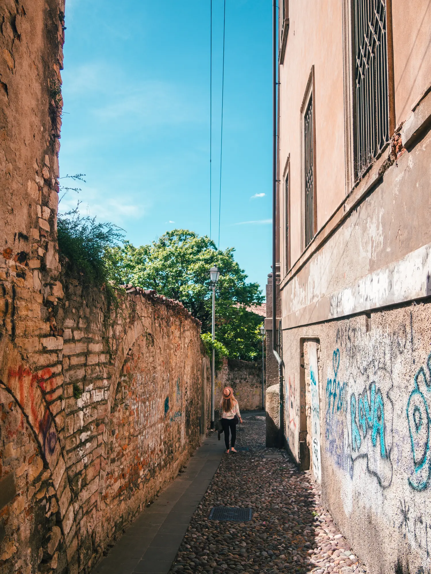 Woman with long hair, wearing black pants, white sweater and a beige hat, walking down a cobbled alleys with beige buildings on either side with graffiti on a sunny day in Bergamo.