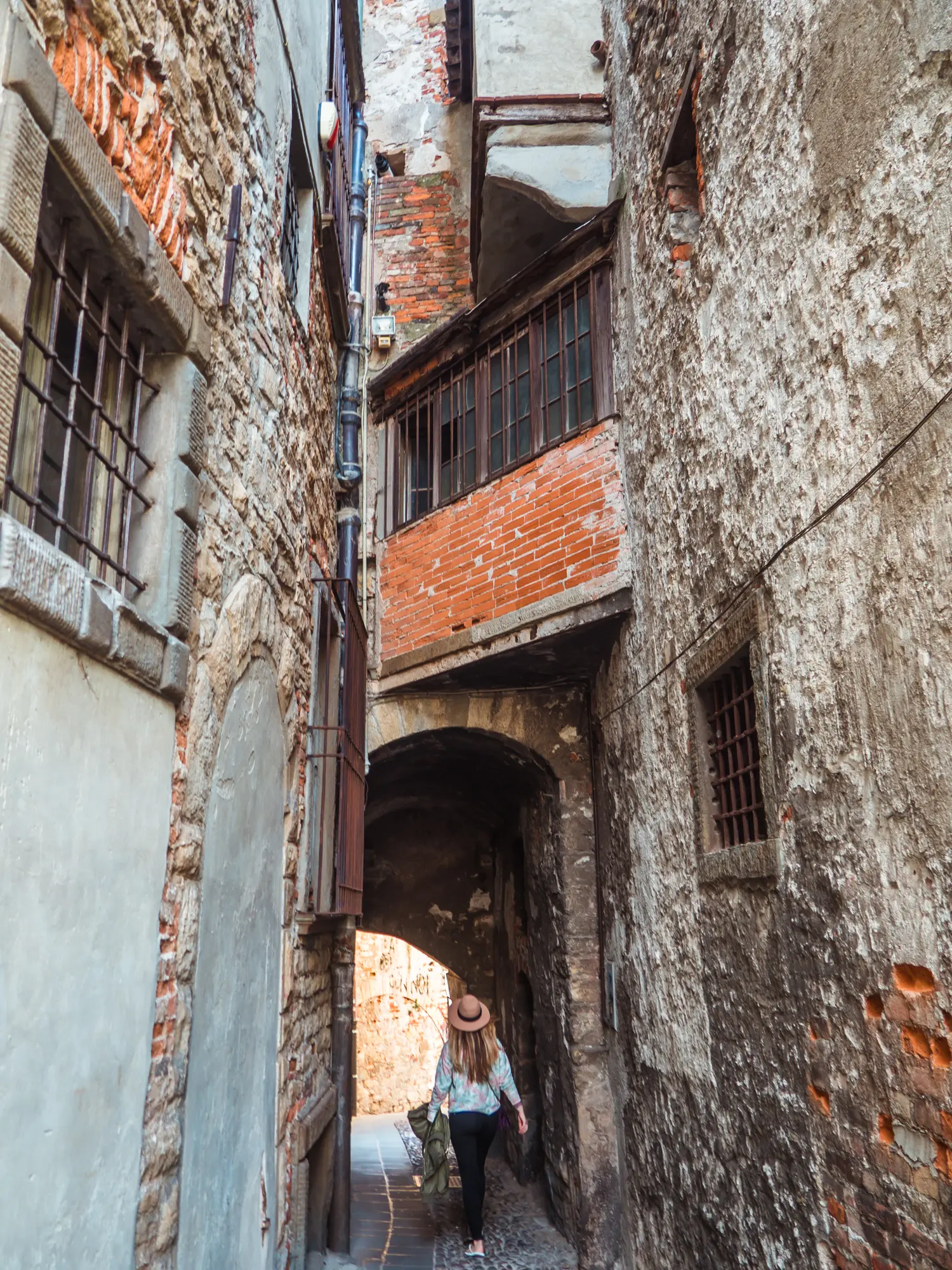 Woman with long hair, wearing black pants, white sweater and a beige hat, walking through a narrow arch with houses built above it on a narrow street in Bergamo, Italy.