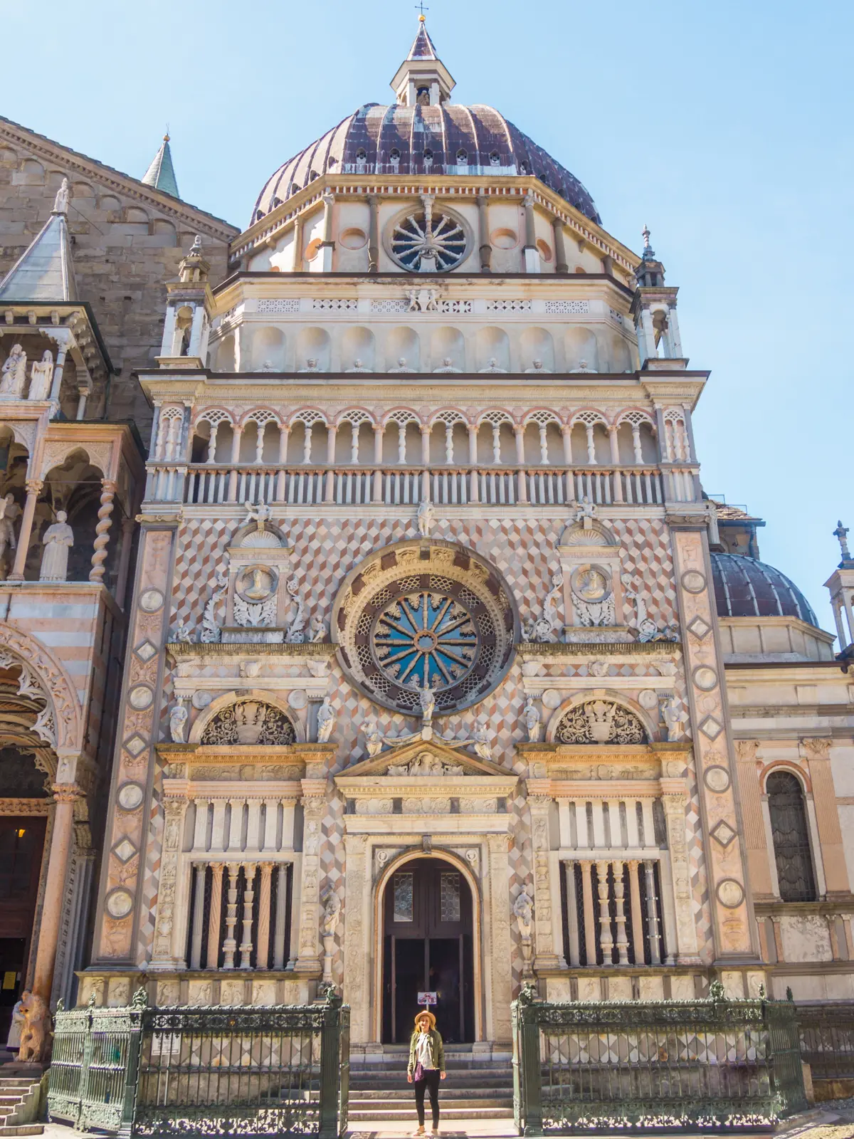 Woman standing between the green gates of Basilica di Santa Maria Maggiore, in front of the ornate facade with a dome, on a sunny day in Bergamo.
