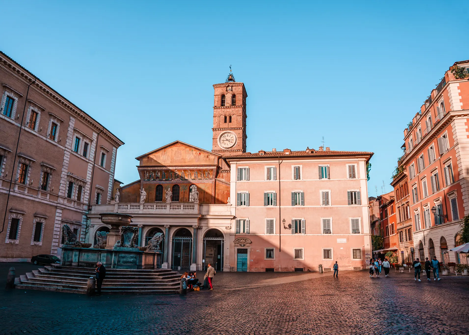 Piazza di Santa Maria in Trastavere, a cobbled square surrounded by light pink and orange buildings on a sunny day, one of the best areas to stay in Rome.