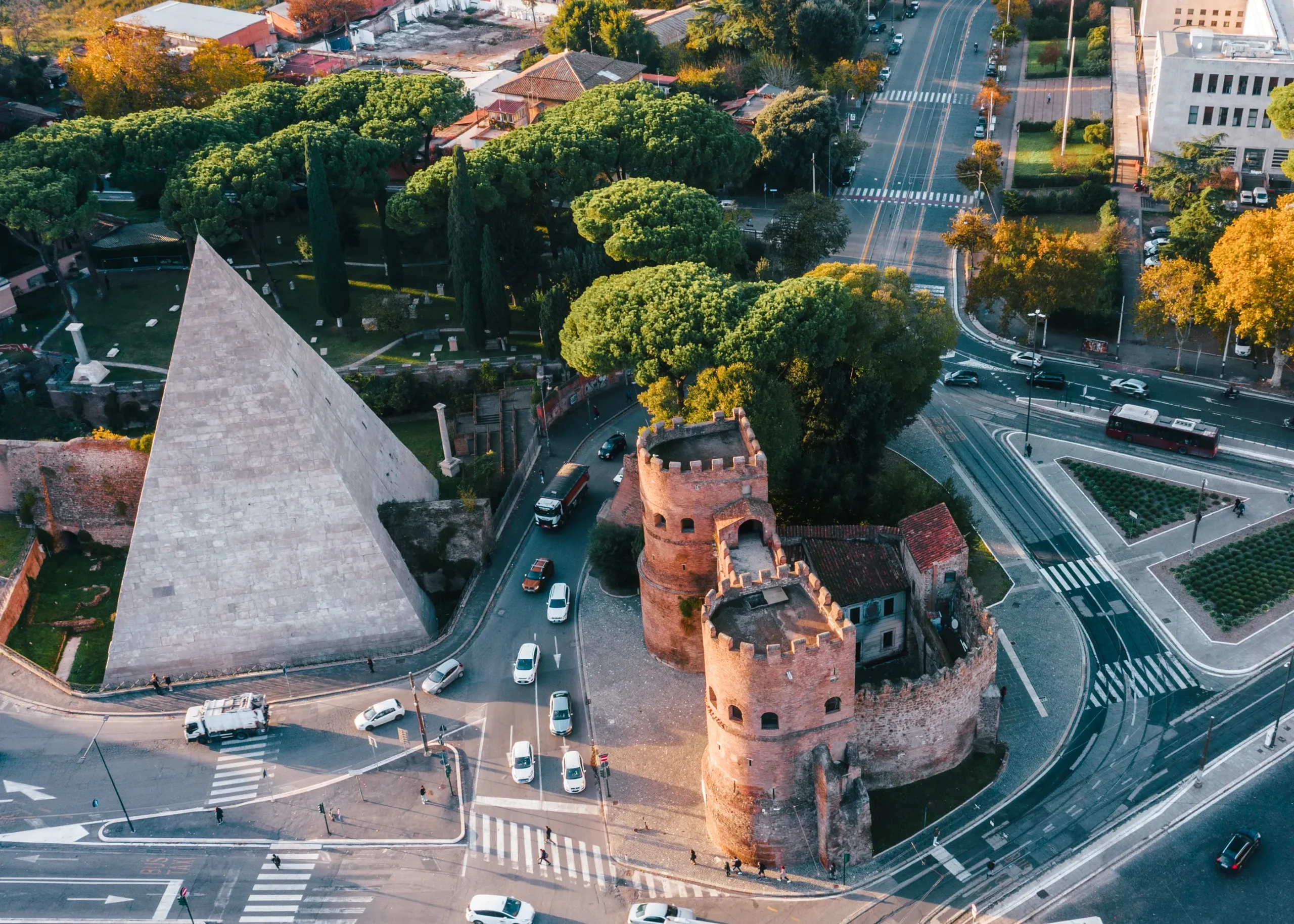 Aerial view of a light grey pyramid and a light brown stone fort surrounded by roads and green trees in the Testaccio neighborhood in Rome, Italy.