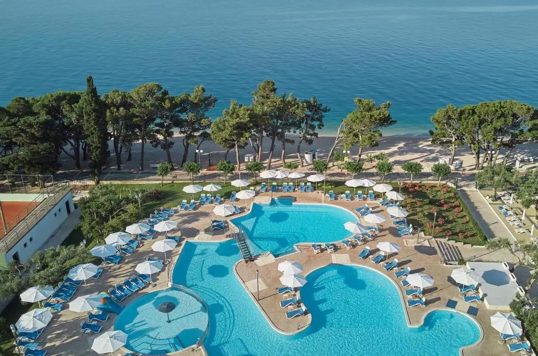 Organically shaped turquoise pool seen from above surrounded by sunbeams and white parasols, set on a pebble beach lined by green trees at Bluesun Hotel Neptun all-inclusive hotel in Croatia.