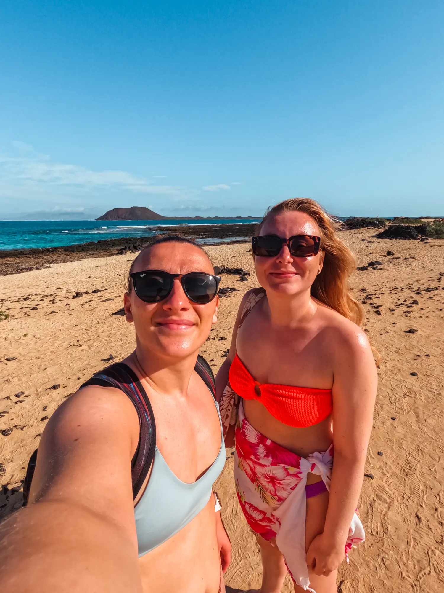 Woman wearing a light blue bikini taking a selfie with another woman wearing an orange bikini top and white sarong on a golden beach on a sunny day in Fuerteventura.