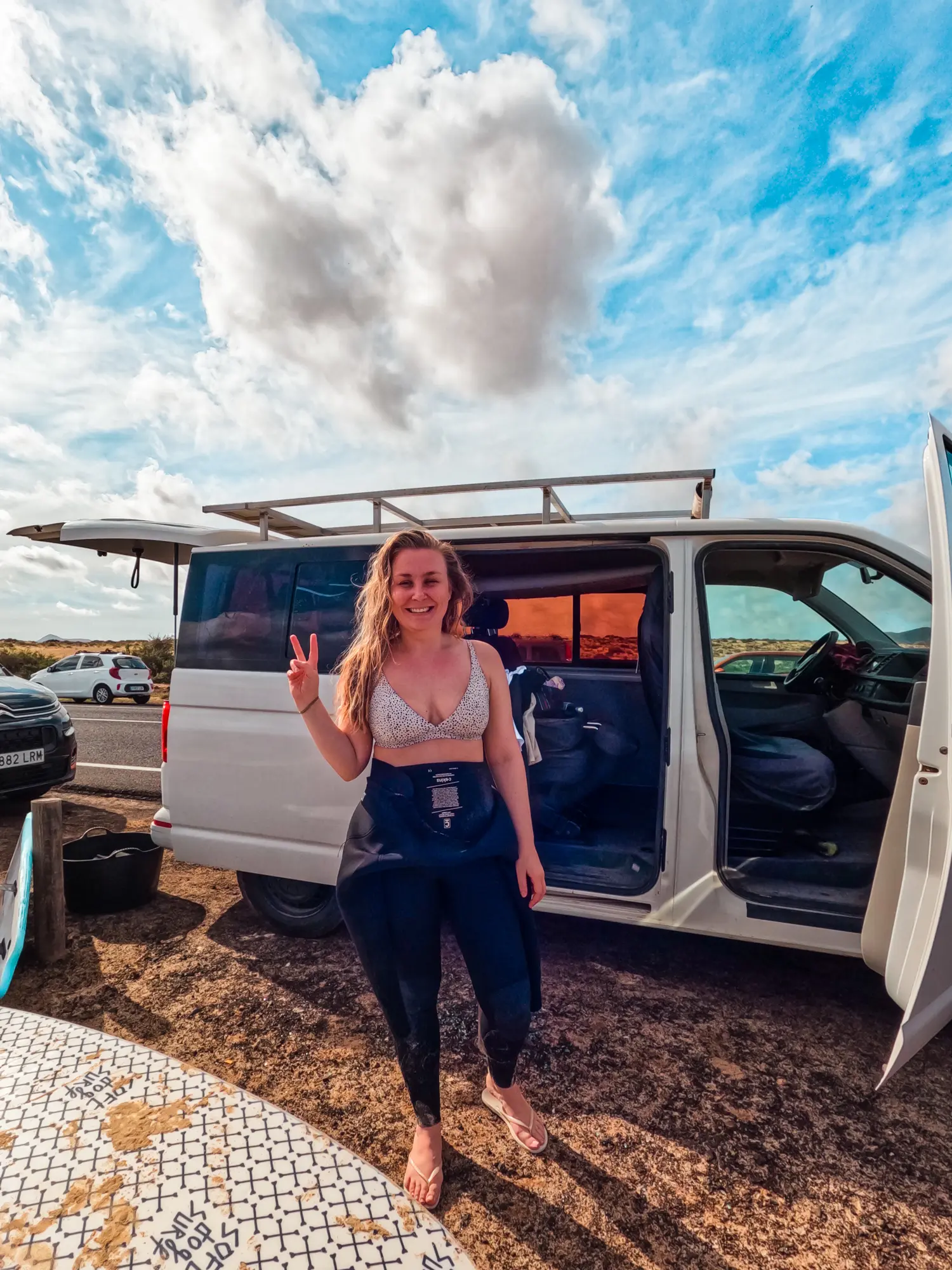 Woman wearing a wetsuit and white bikini showing peace sign in front of a surf school van with doors open in Corralejo Fuerteventura.