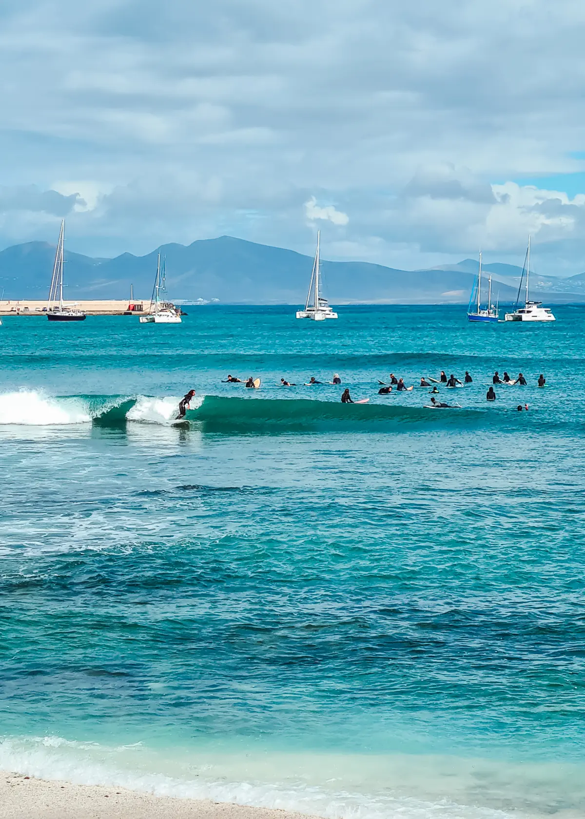 Surfers on a small wave with 20+ surfers sitting in the background in the turquoise ocean on a cloudy day in Fuerteventura.