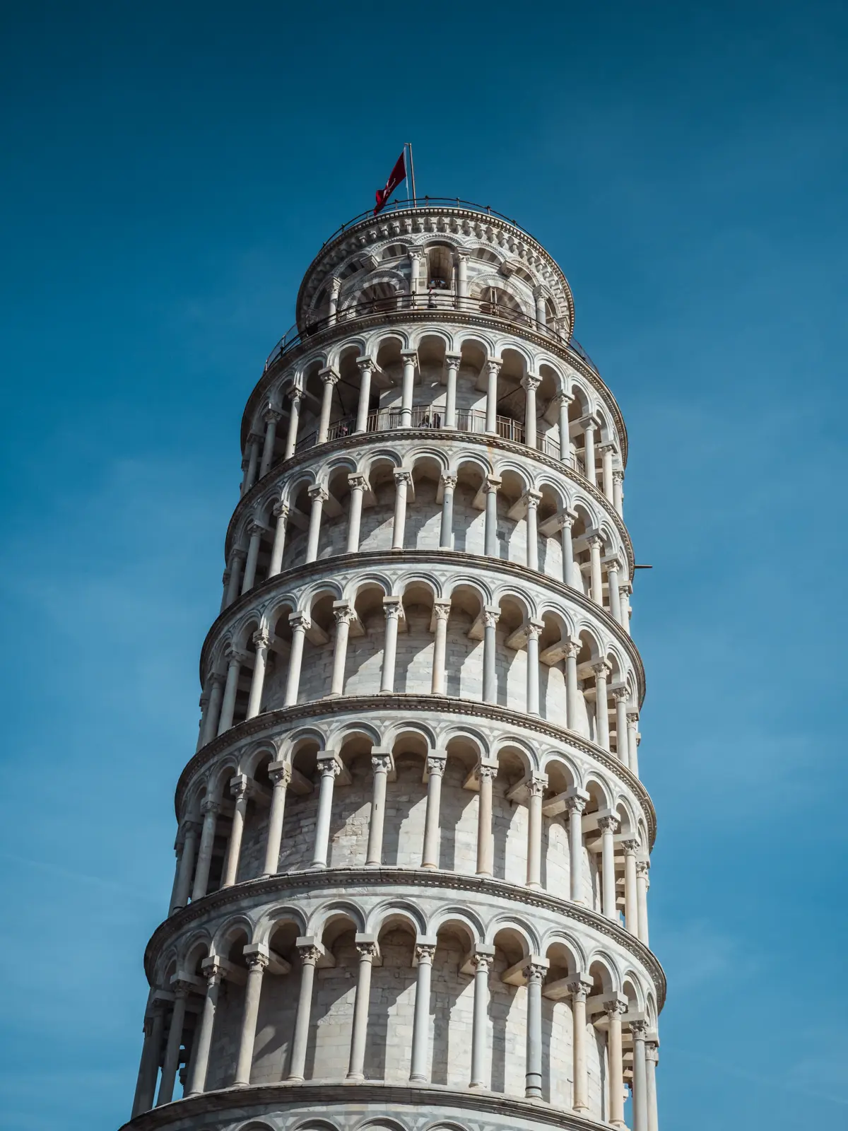 Close up of the Leaning Tower of Pisa with many arches made from white marble and limestone with a red flag on top, set against a deep blue sky.