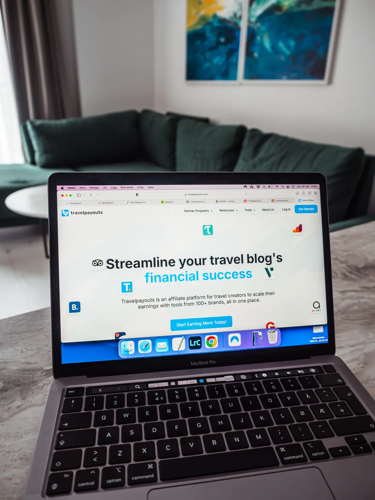 Laptop on a grey table in a living room with blue and green art on the wall, showing the Travelpayouts website. Travelpayouts Review.