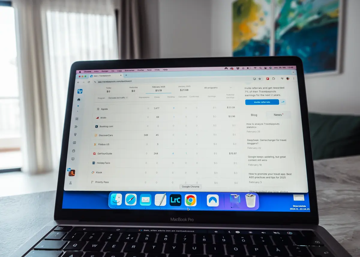 View of the white Travelpayouts dashboard on a laptop on a grey table in a living room with blue and green art on the wall and white curtains on a slider door. Travelpayouts Review.