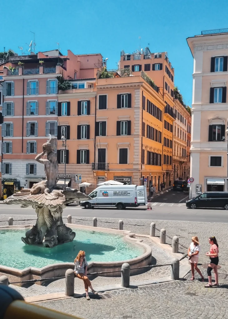 Woman sitting on the Triton Fountain in the Historic Centre with light blue water, and peach and orange colored buildings in the background, one of the best places to stay in Rome.