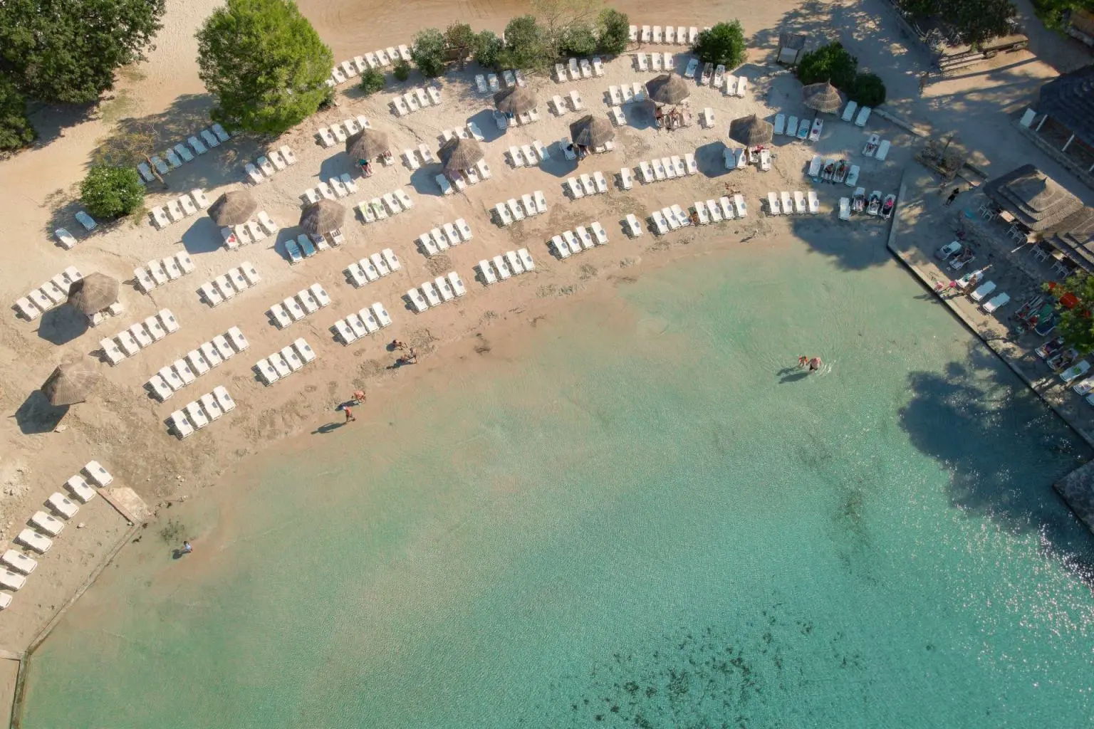 Aerial view of a beautiful light sand beach with rows of white sunbeams and green trees by shallow turquoise water at Pine Beach Pakostane, All-inclusive Croatia.