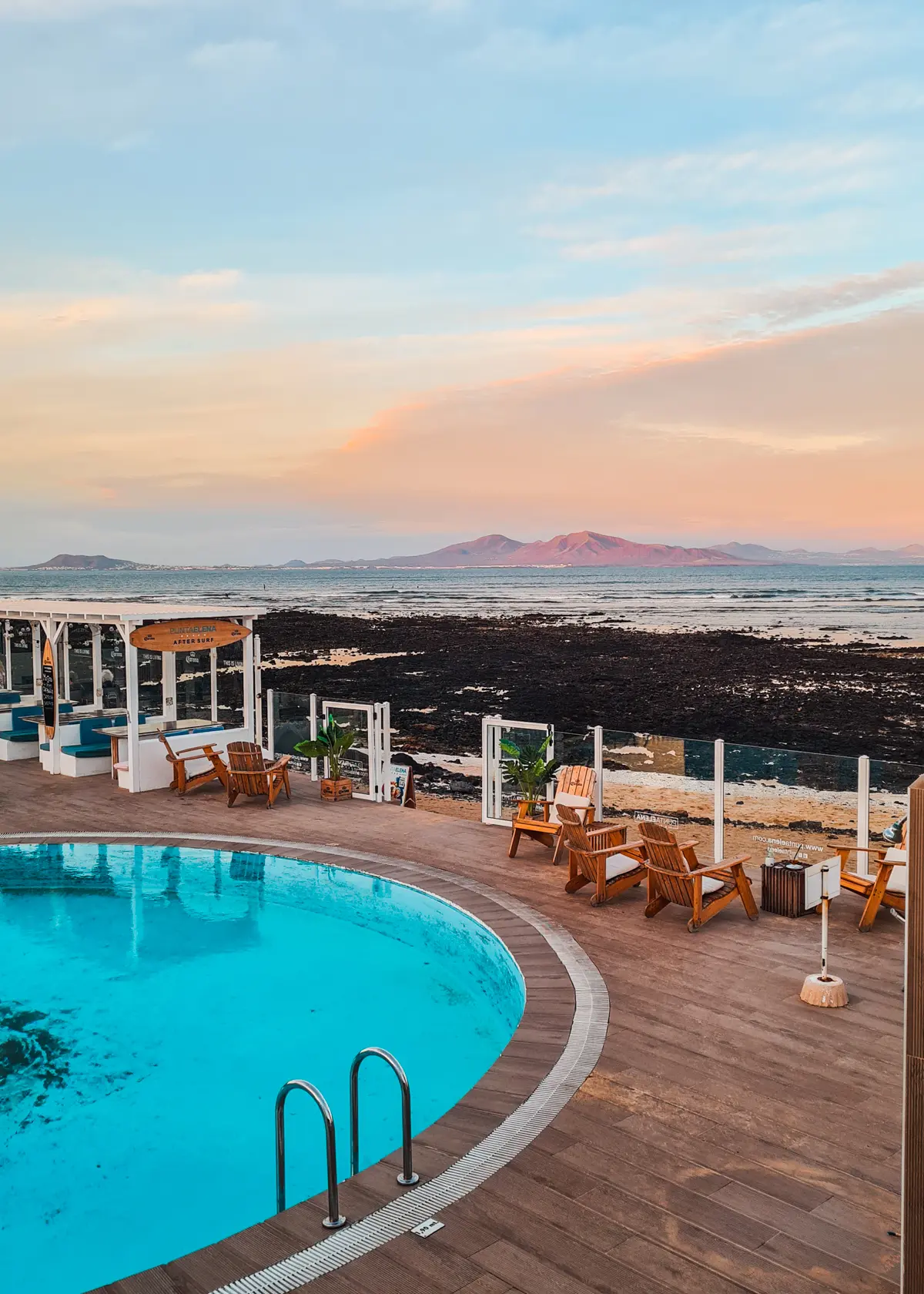 Seaside apartments with a round blue swimming pool, surrounded by wooden lounge chairs and a glass barrier overlooking the ocean, in Corralejo, Fuerteventura. Mountains and and pastel orange sunset in a distance.