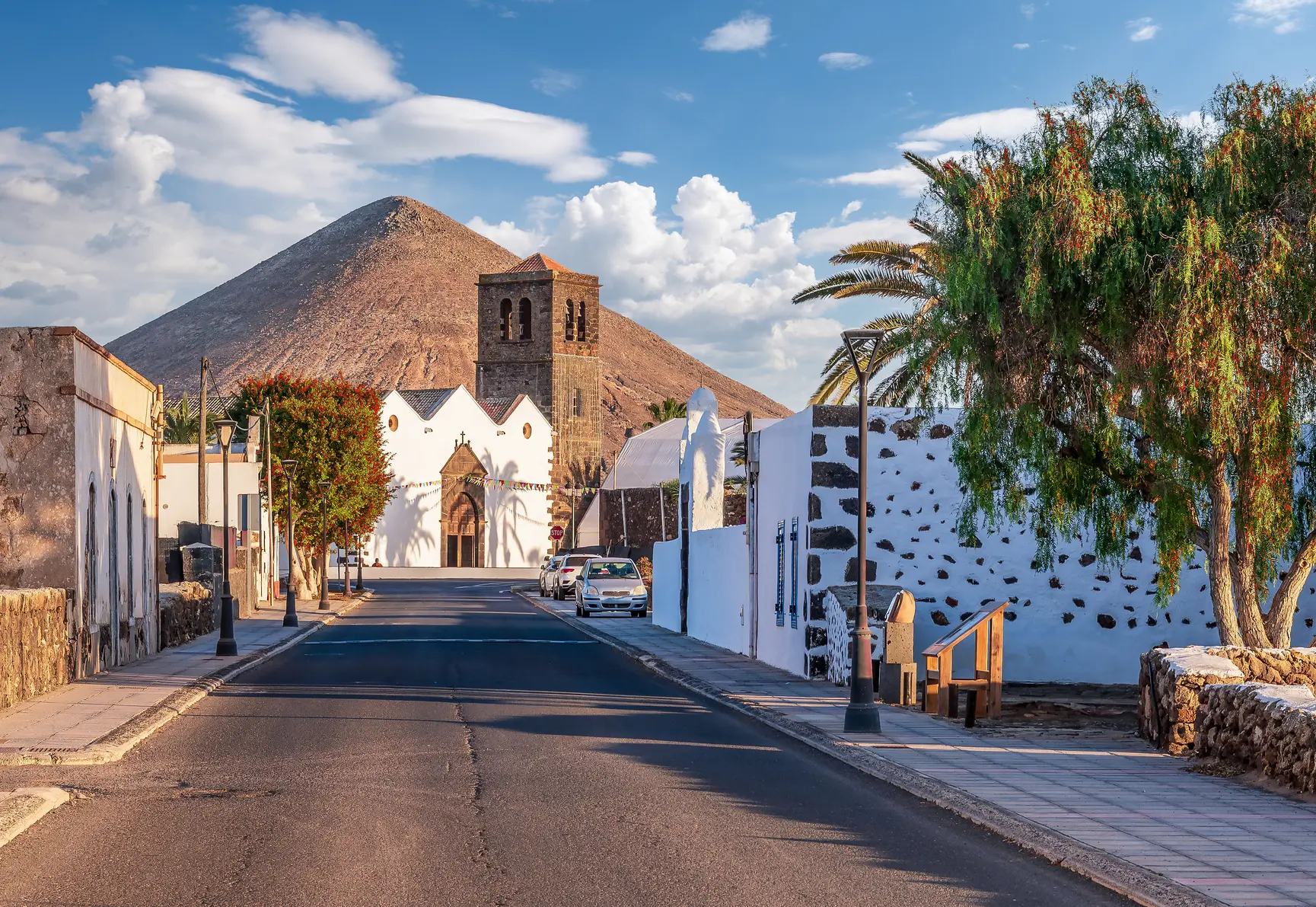 Street leading to a small white church with a brown tower and a dirt hill in the background in La Oliva, Fuerteventura.