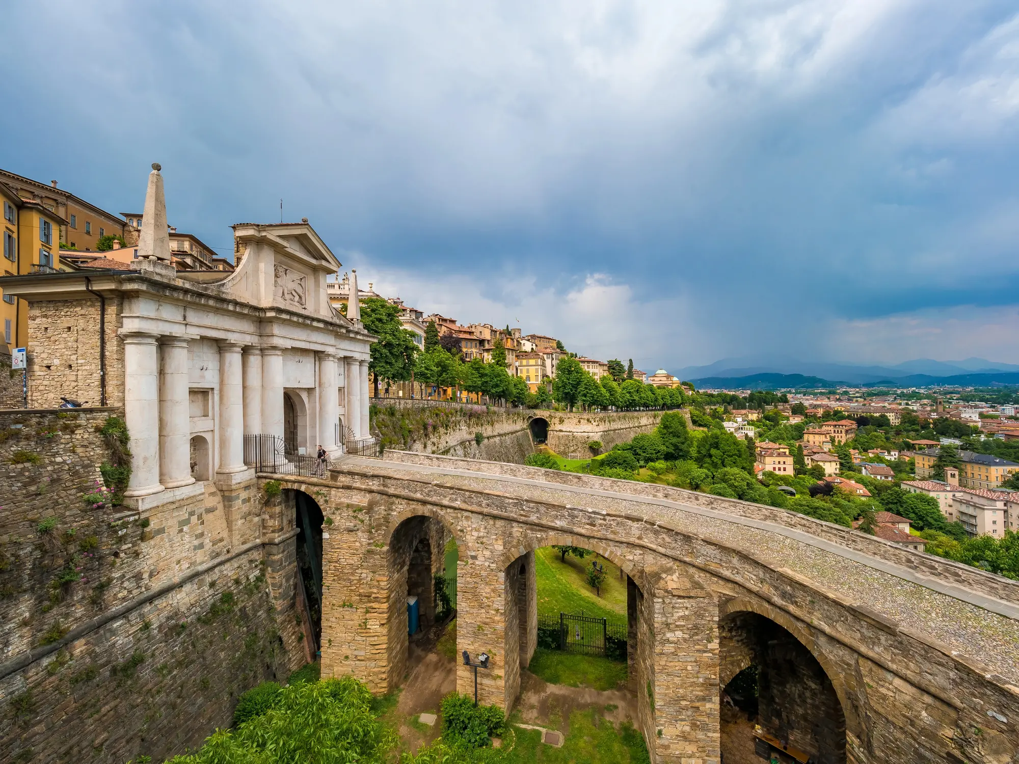 Old stone bridge leading to a large white gate with pillars San Giacomo Gate, with Bergamo Old Town in the background on a cloudy day.