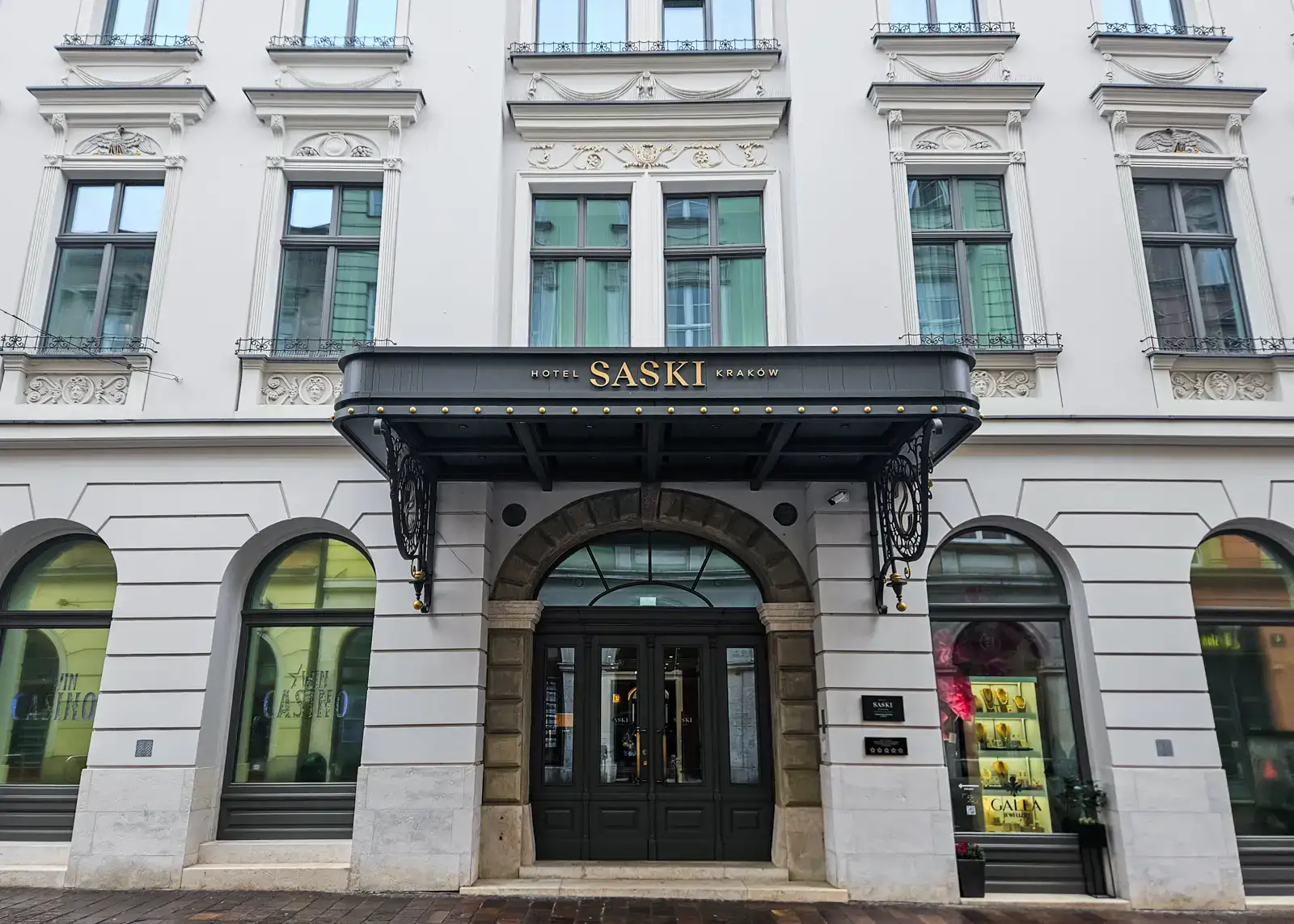 View of the main entrance of Hotel Saki in Krakow in an old, white apartment building with arched windows on the first floor, a black marquee with Saki in gold letters. One of the best places to stay in Krakow.