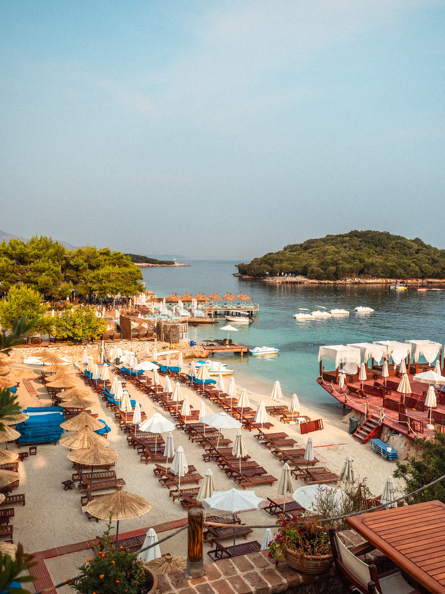 Rows of brown wood sunbeds with white and straw umbrellas with a red deck built over the water at one of the most iconic beaches in Ksamil, with a small green islet in the distance.