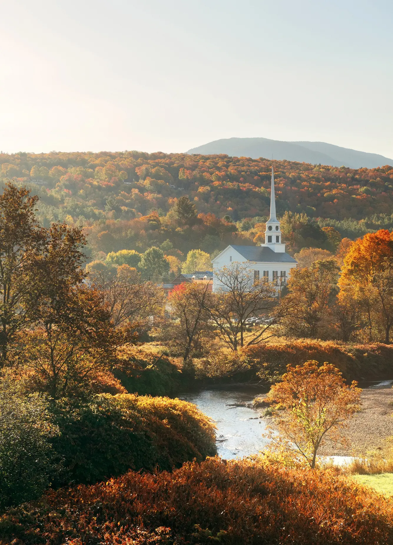 View of a stream in a fall foliage scenery with the white Stowe Community Church with one spire in the morning light, during 3 days in Stowe Vermont.